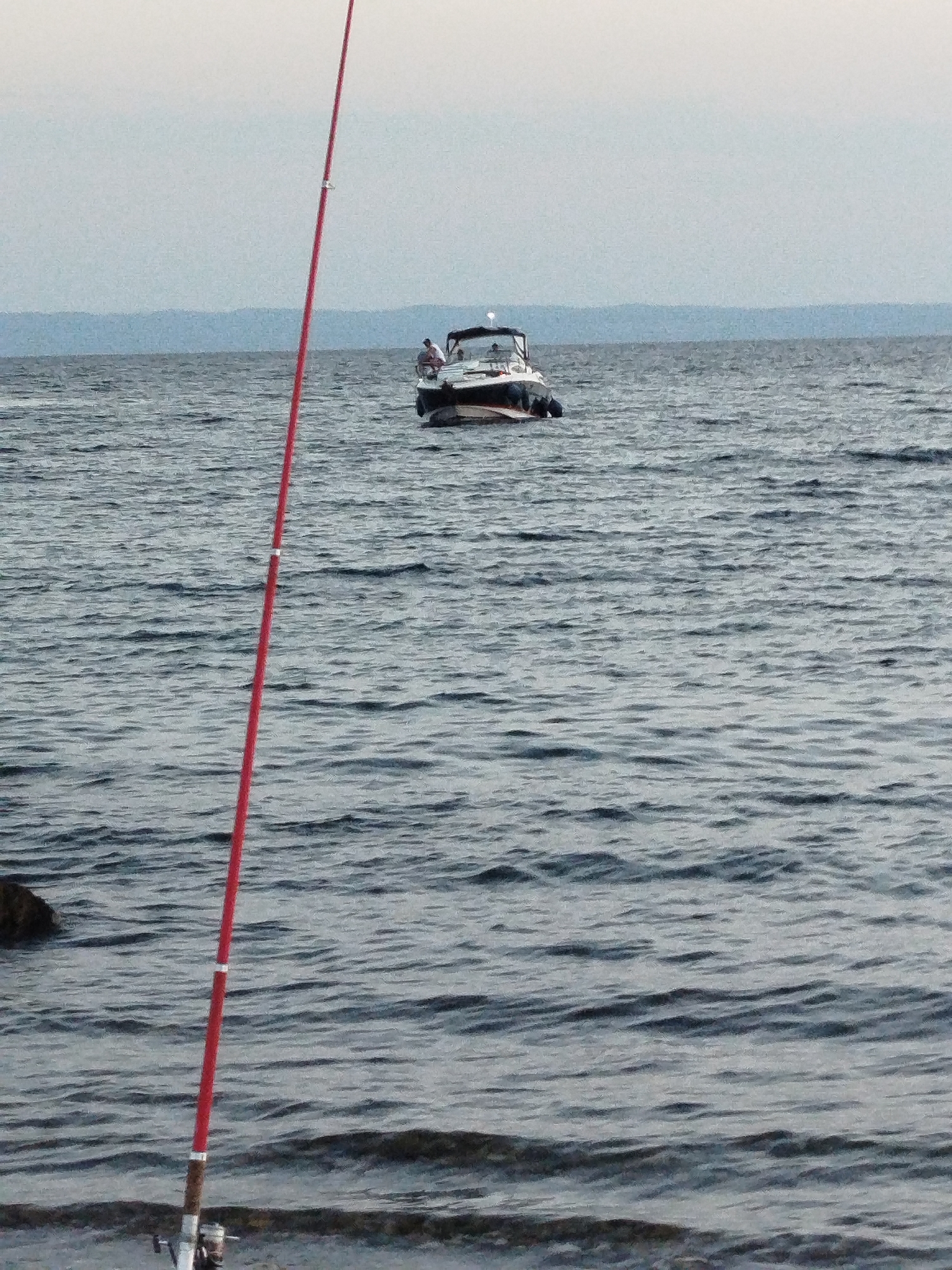The image shows a serene body of water with a fishing rod prominently in the foreground, extending diagonally across the frame from the bottom left towards the top. The fishing rod is red with silver accents. In the mid-ground, a white boat with a dark canopy is anchored or drifting on the water. Two individuals are visible on the boat, one seated and the other standing, both appearing to be engaged in leisure activities. The background features a hazy, blueish horizon line, suggesting distant land or mountains under an overcast sky. The water is choppy with visible ripples and waves, reflecting the muted light. The scene appears to be a calm day at sea, possibly near the coast of Sithonia Municipal Unit, Greece, given the overall landscape and clear water typical of the region.