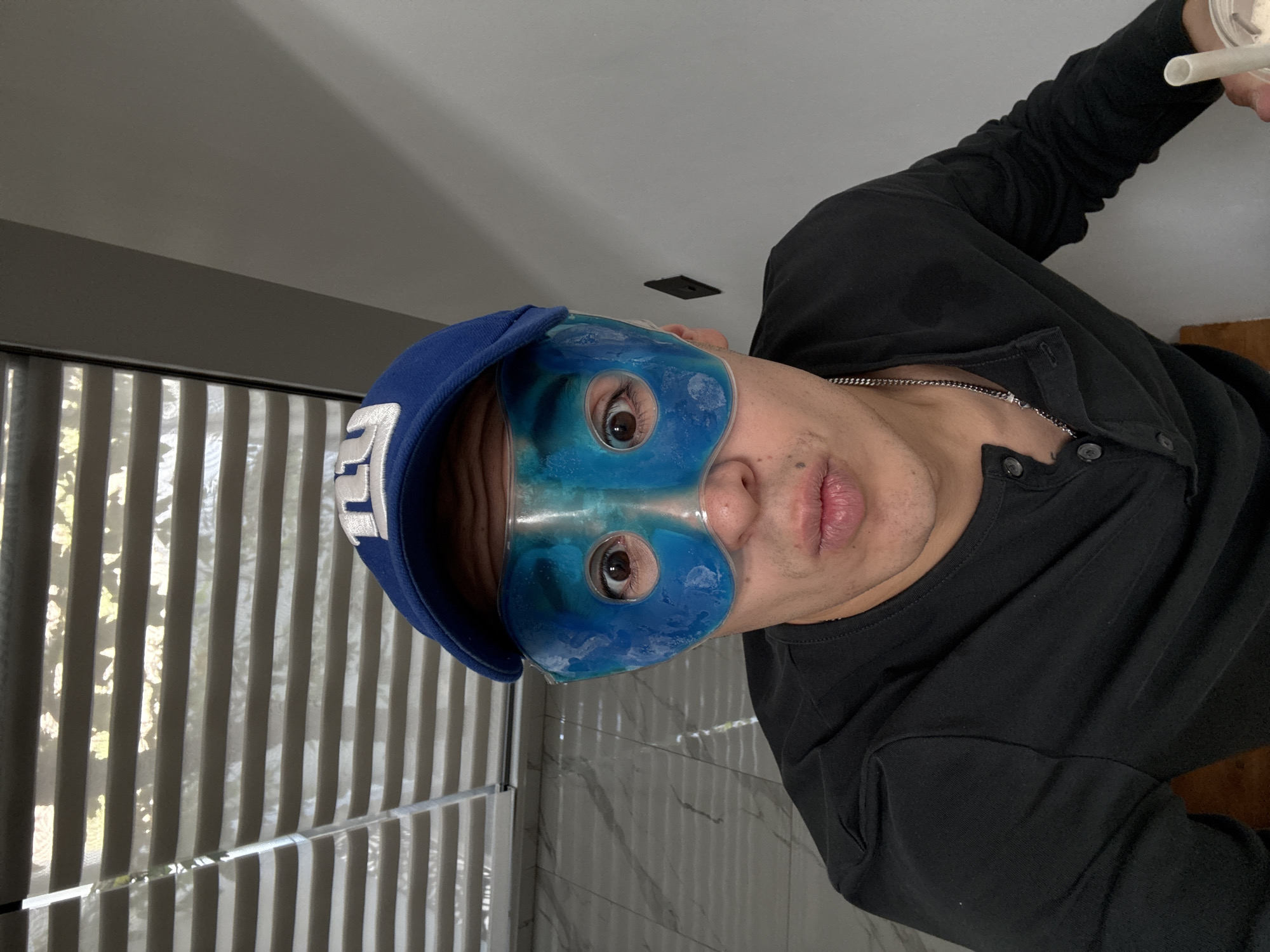 The image captures an adult male indoors, looking directly at the camera. He is wearing a black long-sleeved Henley shirt with buttons at the collar, a silver chain necklace, and a blue baseball cap featuring a stylized white letter "E" (or "EL") embroidered on the front. A translucent blue gel eye mask covers his eyes and upper nose, with his eyes visible through the cutouts. His mouth is slightly pursed. In his right hand, he holds a white cup with a straw.
The background setting indicates an indoor space. To the left, vertical window blinds are partially drawn, allowing natural light to filter in, suggesting it is daytime. The wall behind him is light-colored with a subtle marbled pattern, and a plain ceiling is visible above. The man appears to be engaged in a moment of relaxation or self-care, possibly while drinking from the cup. The location context for the image is San Bernardino Tlaxcalancingo, Mexico.