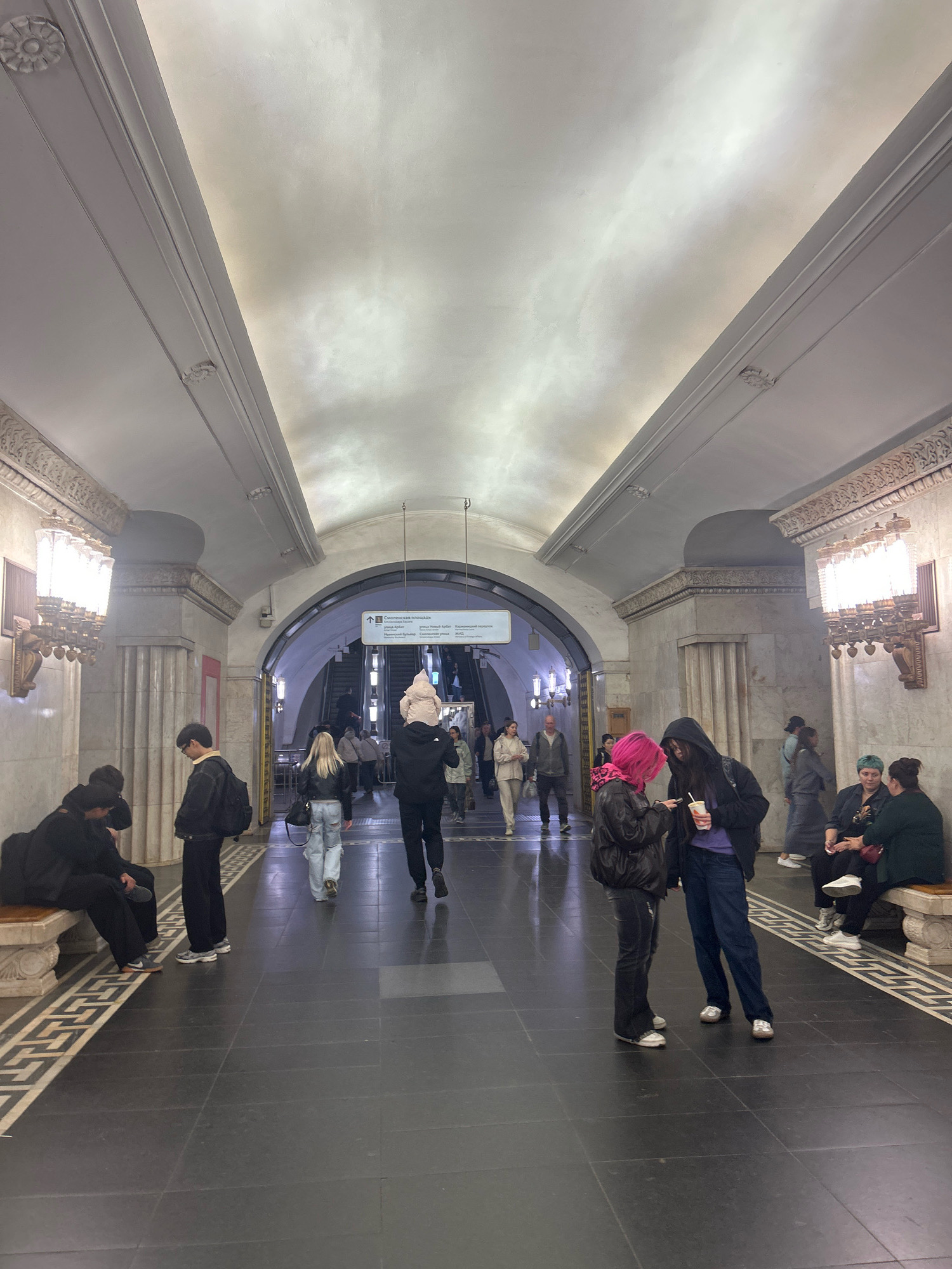 The image depicts an interior scene, likely a subway station due to the presence of escalators, signage, and decorative architecture. The ceiling is high and arched, featuring ornate molding and recessed lighting. Large, decorative lamps with multiple bulbs are mounted on the walls, casting a warm glow. The walls themselves appear to be made of marble or a similar polished stone. In the foreground, people are seen walking and sitting on benches. Some individuals are walking towards an archway leading to escalators, while others are conversing or looking at their phones. The floor is dark and tiled. A sign above the archway indicates directions in a foreign language. The overall impression is one of a grand and well-maintained public space.
