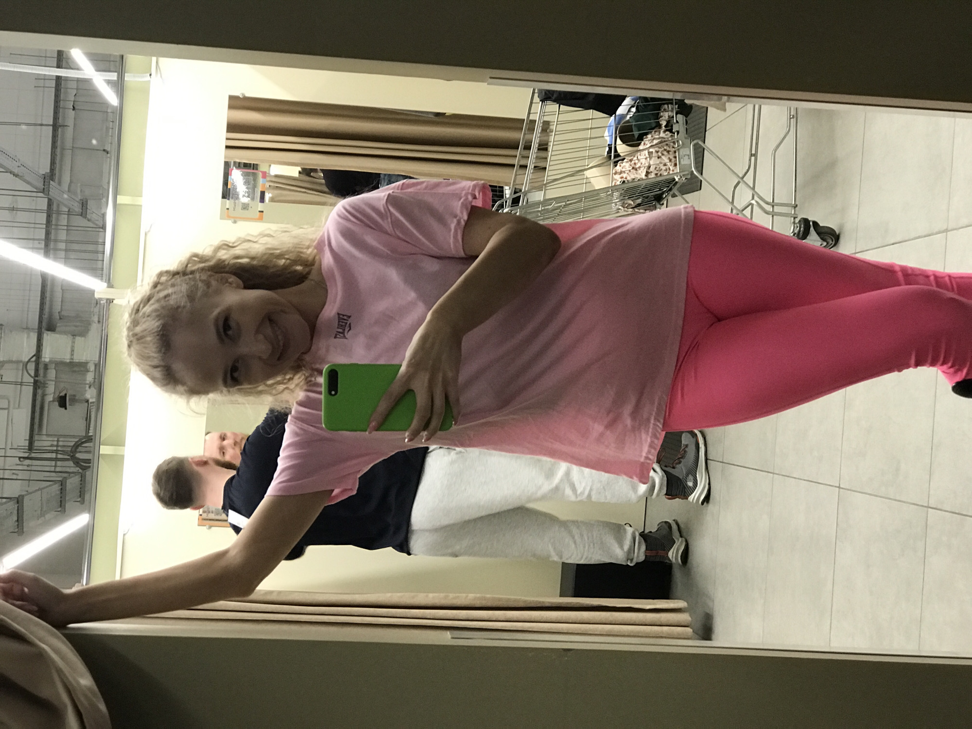 A mirrored image shows a woman with blonde, curly hair holding a green-cased cell phone. She is wearing a pink t-shirt and pink leggings. A man is reflected behind her, wearing a black shirt and light gray pants. A shopping cart filled with items is also visible in the reflection. The location appears to be a retail store in Perm, Russia, based on the tiled floor, clothing racks, and commercial lighting.