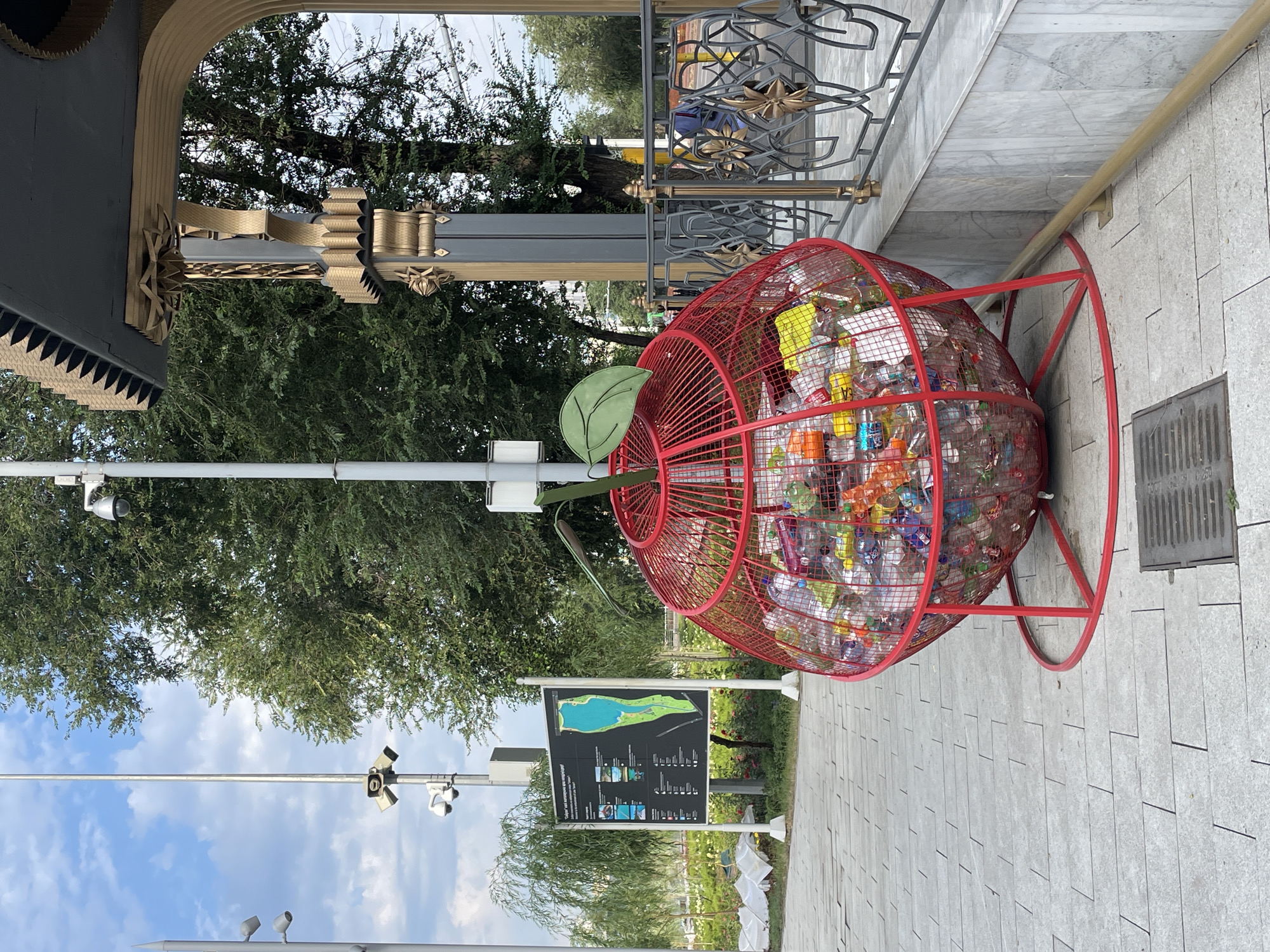 This is a street-level, medium-shot photograph taken outdoors, likely in Almaty, Kazakhstan, given the context provided. The image features a distinctive red, apple-shaped wire trash bin overflowing with plastic bottles. To the left of the bin, a large informational sign is displayed on a white pole, showing a map and text, potentially in Kazakh and Russian, related to a park or recreational area. Behind the sign and bin, lush green trees form the backdrop. In the upper left quadrant, a lamppost with multiple lights and cameras is visible. To the right of the bin, an ornate architectural element with gold accents and a dark gray roof suggests a structure like a pavilion or entrance. A metal railing with decorative patterns separates this area. The ground is paved with light gray rectangular tiles. The sky is partly cloudy with a bright, daytime atmosphere.