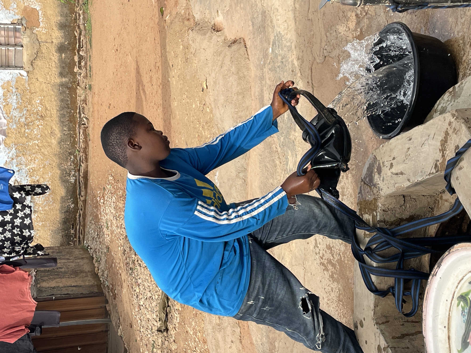 A young man in a blue, long-sleeved shirt with white stripes on the arms and ripped jeans is drawing water from a tap. The water is cascading into a black bucket. The setting appears to be outdoors with concrete walls and ground. In the foreground, a blue and black strap is visible. The location is likely in Jos, Nigeria. Other clothes lie on the ground in the foreground.