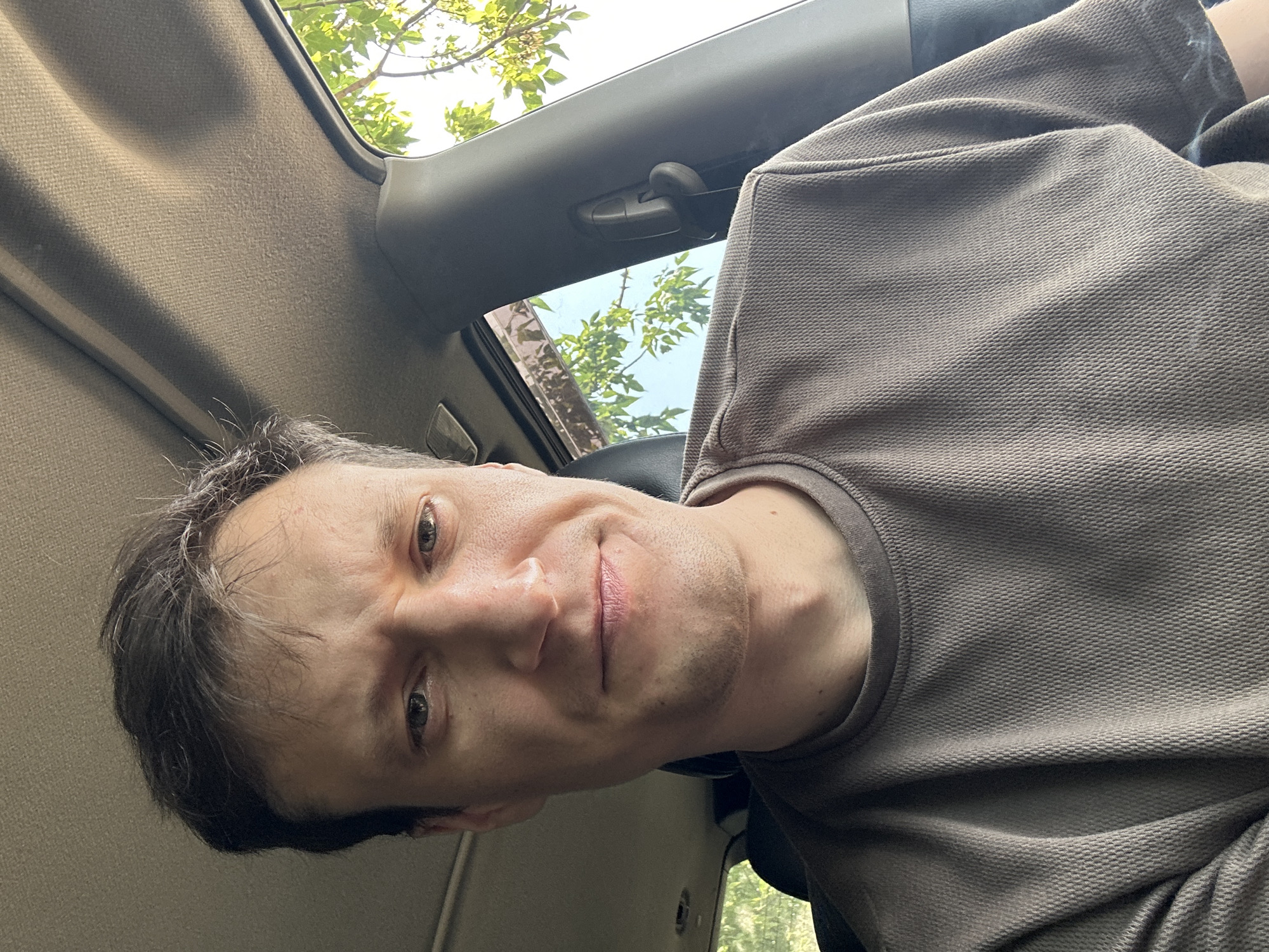 A medium shot, captured from a slightly low angle, shows a man from the chest up, looking directly at the camera. He is seated inside a vehicle, likely a car, given the interior view. The man has light brown hair, styled casually with some strands falling onto his forehead. His facial expression appears slightly concerned or questioning, with his eyebrows furrowed. He has a fair complexion and is wearing a textured, taupe-colored t-shirt.
The background reveals the interior of the car, with a portion of the headliner visible at the top of the frame and a dark seat behind him. To the right, a car pillar and a part of a seatbelt mechanism are visible. Sunlight streams in through a window on the right side, illuminating the scene and casting soft shadows. Through the window, glimpses of green foliage from trees and a bright blue sky suggest an outdoor environment, possibly during the day.
The location is identified as Makiv, Ukraine, implying this image was taken within or near this Ukrainian locality. The overall impression is of a candid self-portrait taken by the man while inside his vehicle.