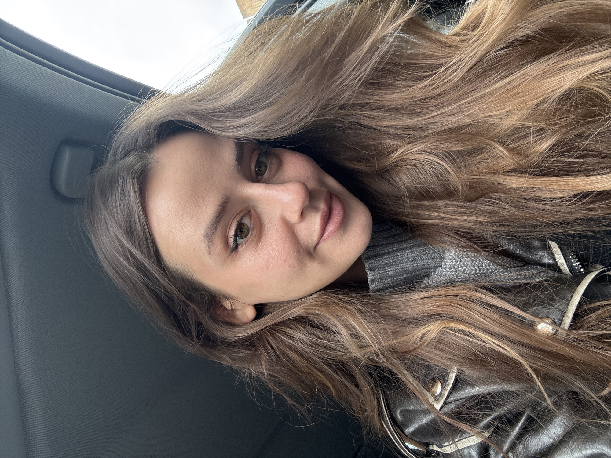 A young woman with long, wavy brown hair poses for a selfie. She is fair-skinned with light brown eyes. She is wearing a gray sweater and a black leather jacket. The background is a car interior, with the car's ceiling visible in the upper left corner. The photo was taken indoors. It is unknown if it was taken in Krasnoyarsk, Russia.