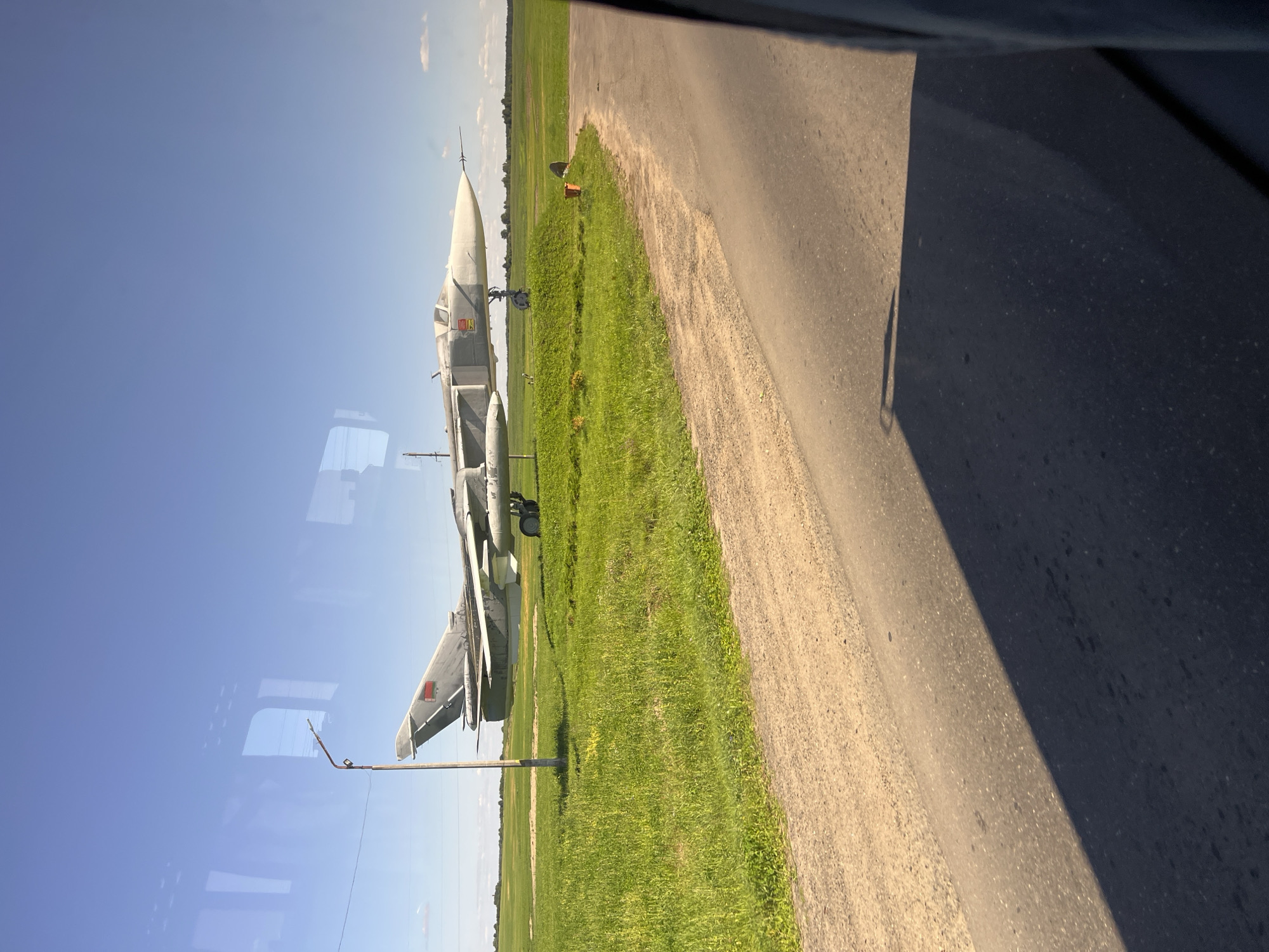 The image depicts a military jet, likely a Sukhoi Su-24, displayed on a grassy field under a clear blue sky. The aircraft is predominantly grey with some markings, including a red star symbol on its tail fin, suggesting its origin. It is positioned on a landing gear, indicating it's not in flight. The setting appears to be an open area, possibly a museum, park, or a historical display, given its static placement. Surrounding the jet is a vibrant green field with some scattered vegetation. A dirt road and a paved asphalt road are visible in the foreground, with shadows cast across the asphalt, implying a sunny day. A lamppost stands to the left of the aircraft. Reflections of windows are visible on the left side of the image, hinting that the photo was taken from inside a vehicle or building with large windows. The environment suggests a rural or semi-rural location. Based on publicly available information, a Sukhoi Su-24 fighter-bomber is displayed at the Museum of Traditional Folk Crafts and Local Lore in Pastavy, Belarus. This display likely commemorates military history.