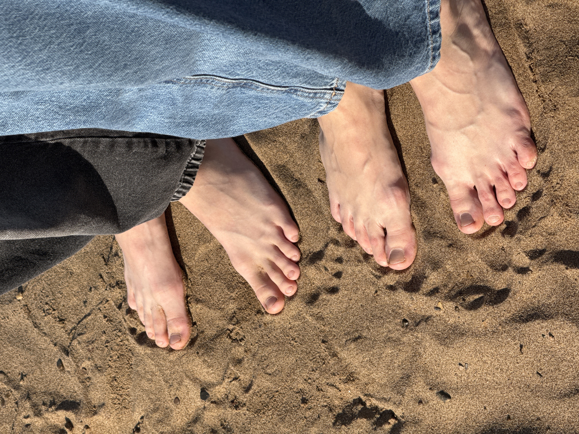 The image depicts four feet standing on a sandy surface. Two of the feet belong to a person wearing light-wash denim jeans, and the other two belong to a person wearing dark-wash denim jeans. The sand is brown and contains small pebbles. There are footprints visible in the sand. The image suggests a location near a beach or sand dune, however, there is no evidence that is this in Chaikovsky, Russia.