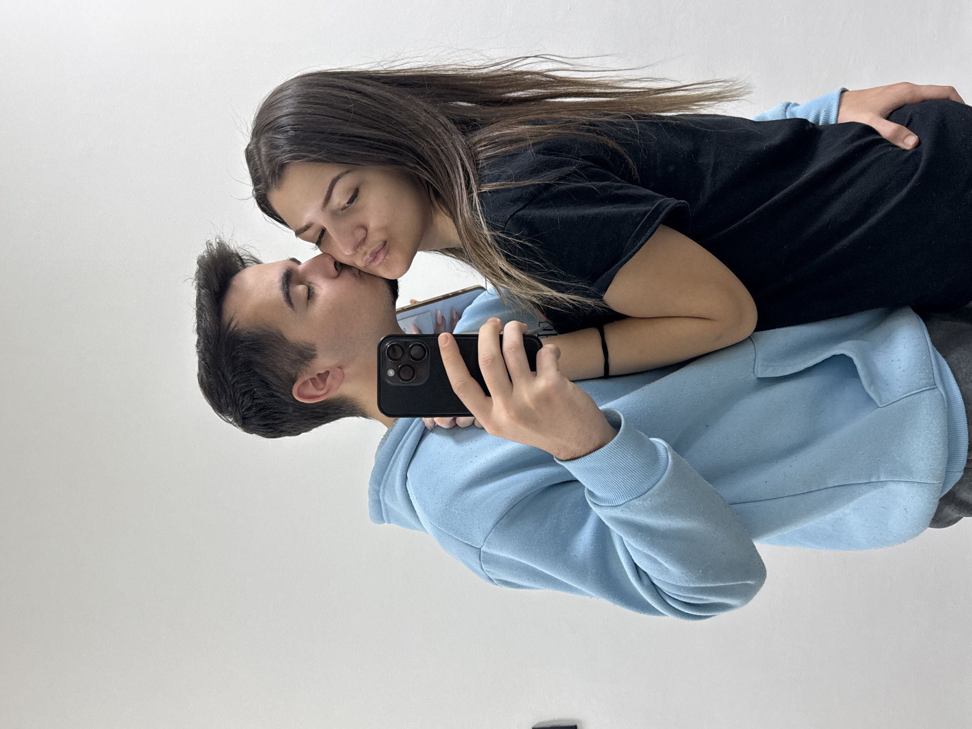 A young couple is posing for a selfie in what appears to be an indoor location, possibly a private residence. The man, wearing a light blue hoodie, is holding a black smartphone, presumably taking the photograph. The woman, with long brown hair and wearing a black shirt, is kissing the man on the cheek. Her left hand is on his waist. The background is a plain white wall. There are no indications of the location being in Sofia, Bulgaria.