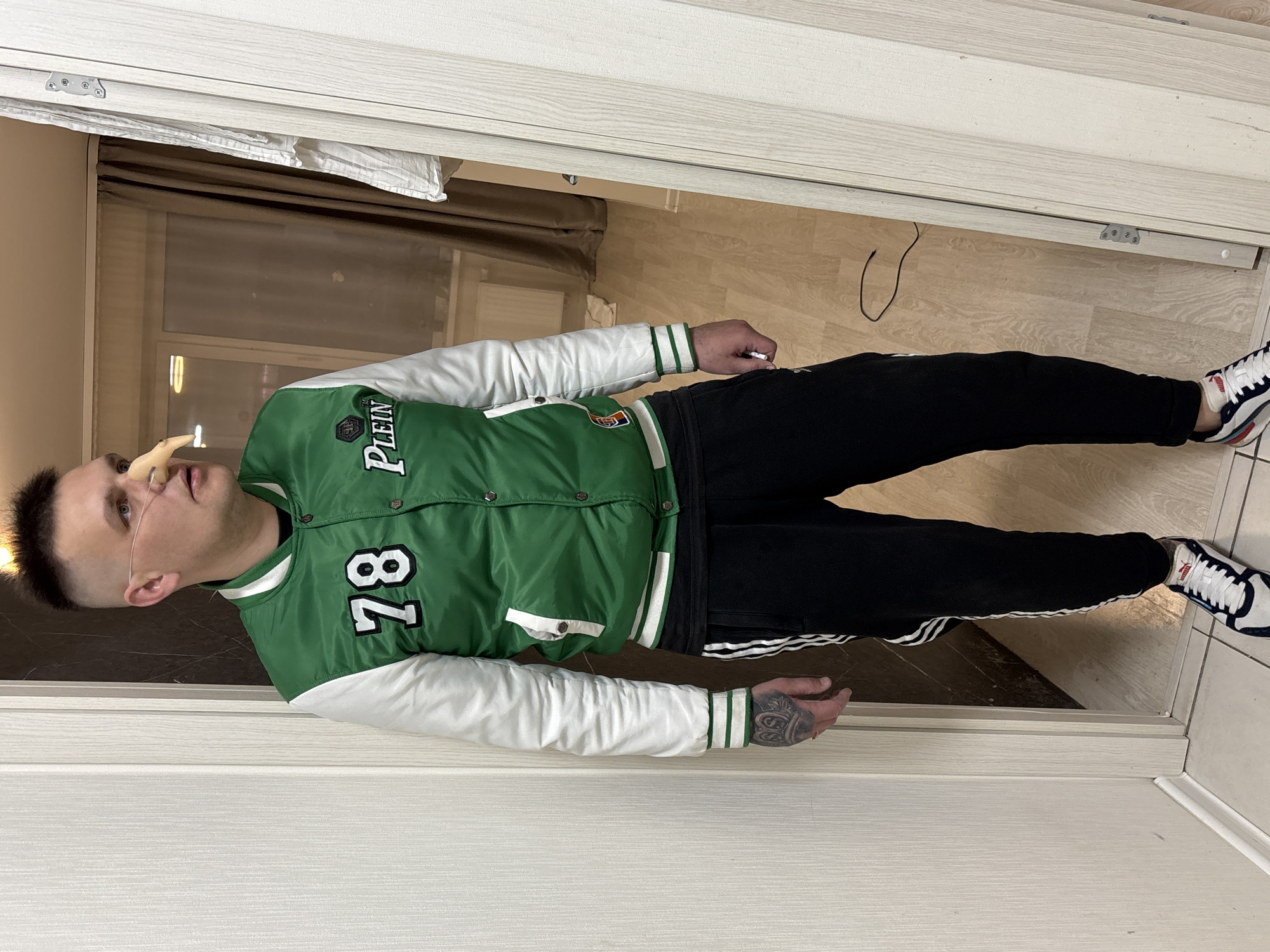 A man wearing a green and white jacket, black pants, and white sneakers stands in a doorway. He is wearing a nasal cannula. He has a tattoo on his right hand. The scene appears to be indoors, with a glimpse of a room visible behind him. Based on visual details of the background and the context provided, it is plausible that the location is in Izhevsk, Russia.