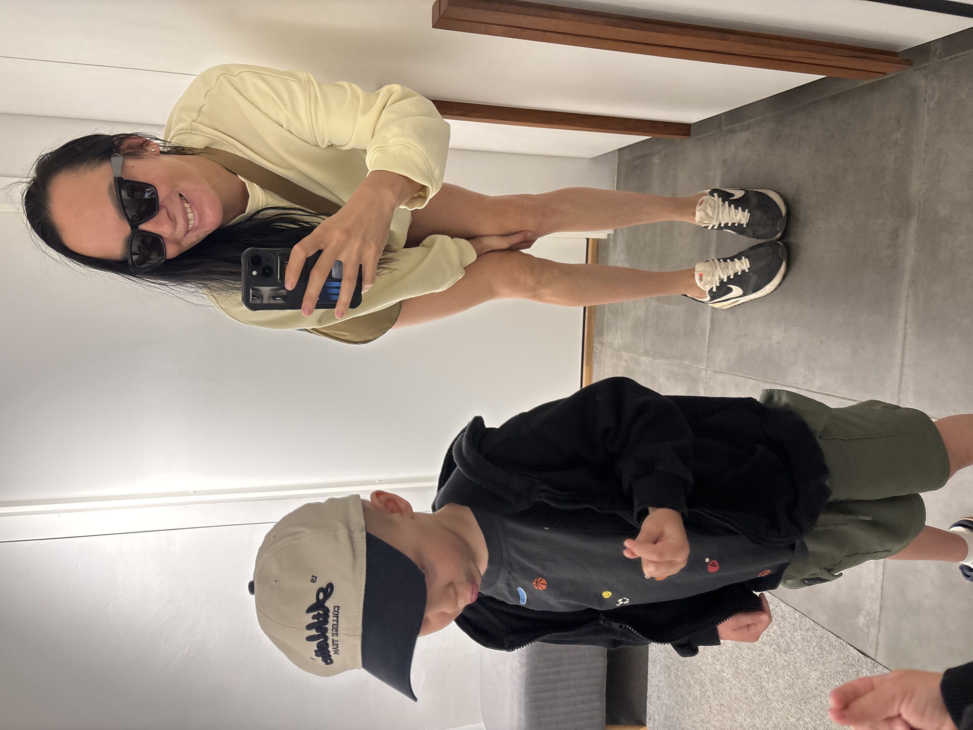 A woman and a young child are in a room. The woman is smiling and appears to be taking a selfie. She is wearing sunglasses and a light yellow sweatshirt dress, along with black and white sneakers. The child is wearing a cap, a gray shirt with small ball patterns, a black hoodie, and green shorts. The floor is light gray and appears to be concrete or tile. A wooden structure is visible on the upper part of the wall. A part of a gray sofa is visible at the bottom of the picture. The photo was taken in Chaikovsky, Russia.
