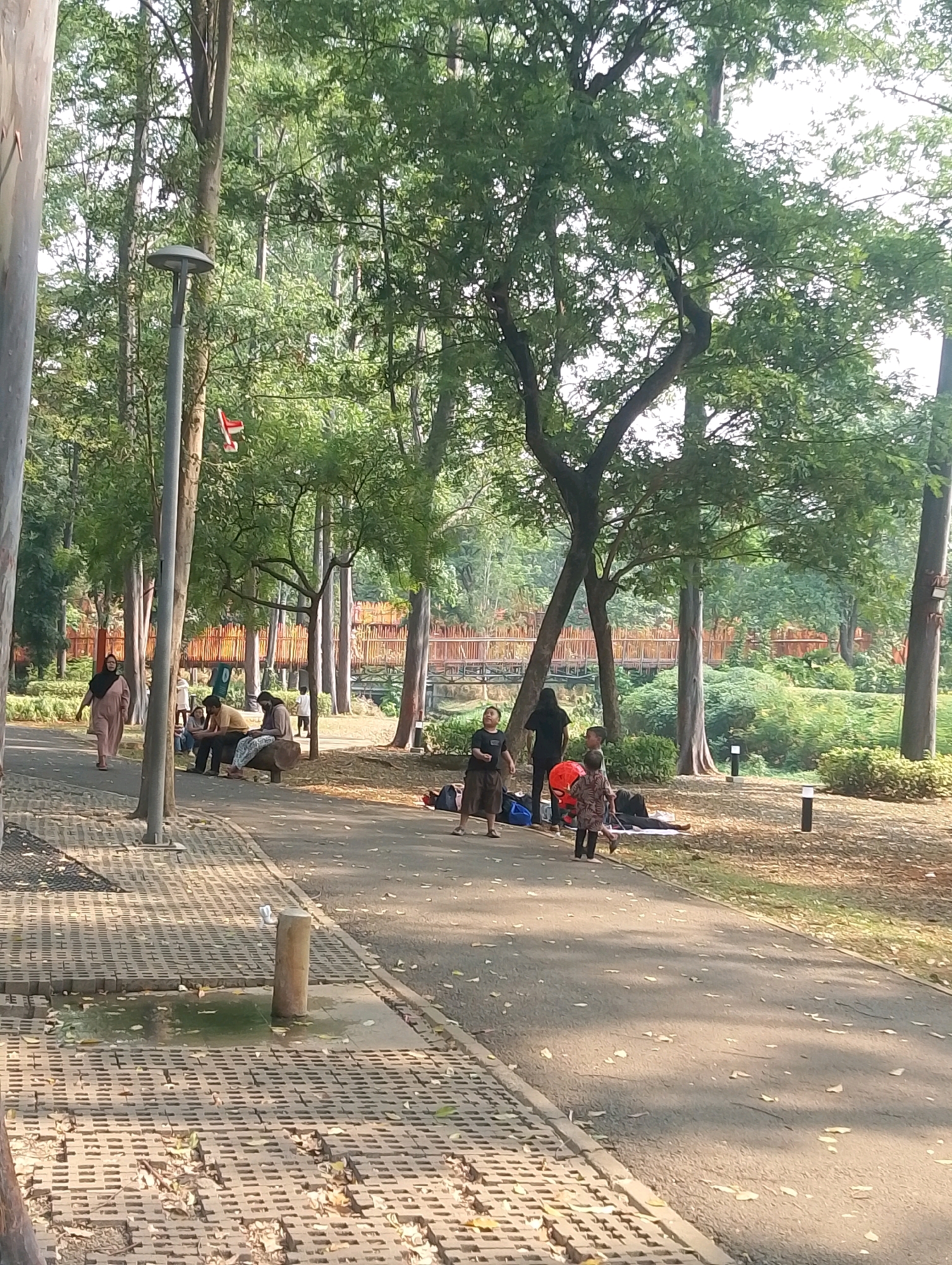 The image depicts an outdoor scene within a park setting, possibly in South Jakarta, Indonesia, given the presence of lush trees and open green spaces. In the foreground, a paved walkway with a patterned brick border runs diagonally across the frame, with a shallow pool of green water visible on the brickwork. Several people are scattered throughout the park.
On the left side of the walkway, a woman in a light pink hijab and dress is walking away from the camera. Further along the path, a group of individuals are seated on a log-like bench. One person is wearing a yellow shirt and another a patterned fabric. Beyond them, a woman in a black hijab and long black garment stands facing away from the viewer.
In the middle ground, the park transitions into a grassy area with more trees. A red, elevated pedestrian bridge with a lattice structure is visible in the background, spanning across a natural-looking divide, potentially a dry creek bed or a lower level of the park. A few people are visible near the bridge, some sitting on the ground.
Several trees with dense green foliage dominate the background, creating a dappled sunlight effect on the path and ground. There are also several tall, thin trees, some with visible bark texture, and a streetlamp with a broad, flat top is positioned on the left side of the path. Small, dark, cylindrical objects resembling outdoor lights are also present in the grassy areas. The overall atmosphere is one of a relaxed, public park on a bright day.