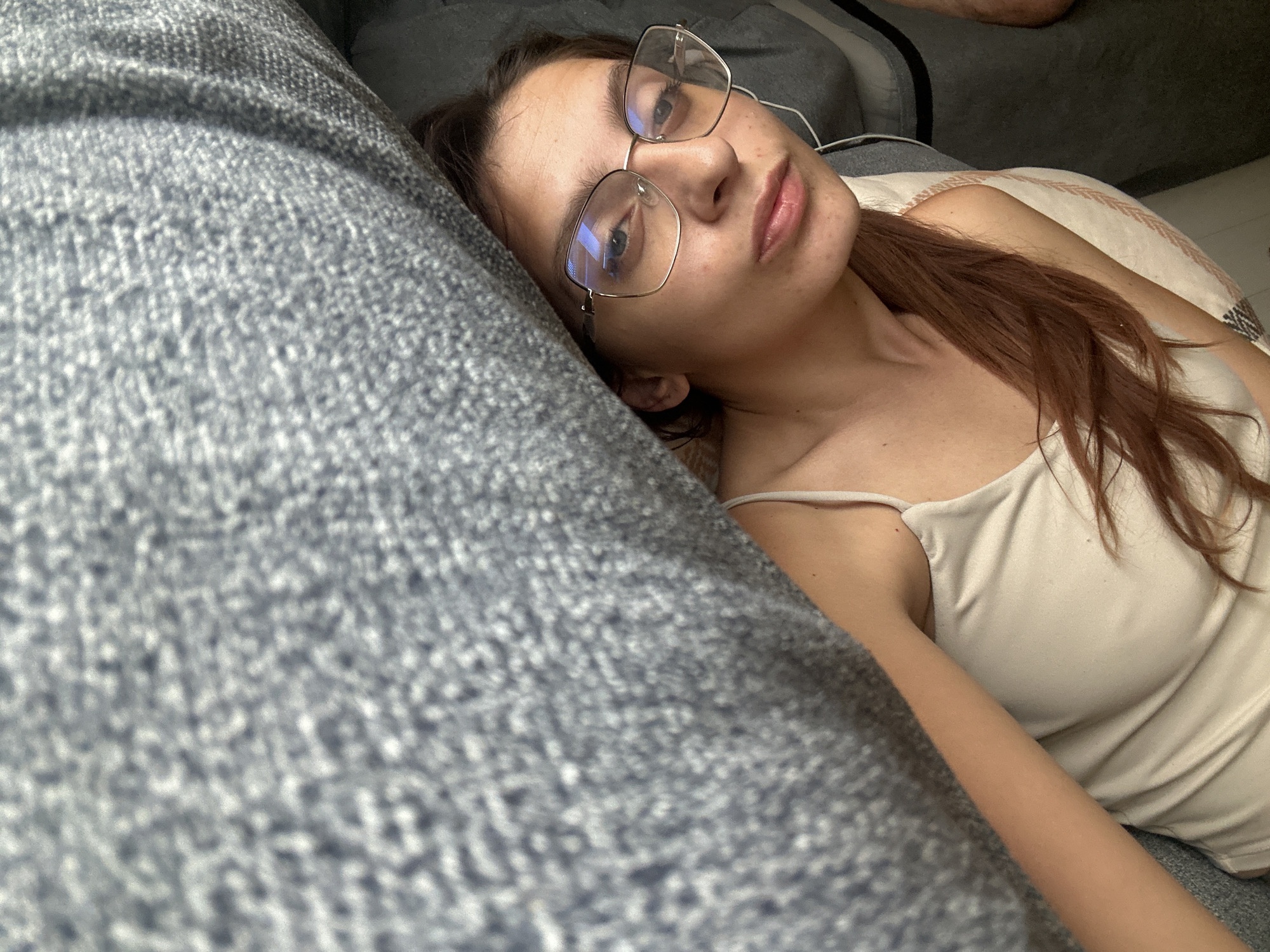 A young woman with long, auburn hair and clear skin, wearing glasses and a light-colored tank top, is lying down on a textured grey sofa. She is positioned on her right side, with her head resting on a pillow. Her gaze is directed towards the camera. The environment appears to be a domestic setting, possibly a living room. There are no identifiable landmarks or clues to pinpoint the location within the city of Tryavna, Bulgaria. The overall scene depicts a casual, relaxed moment.