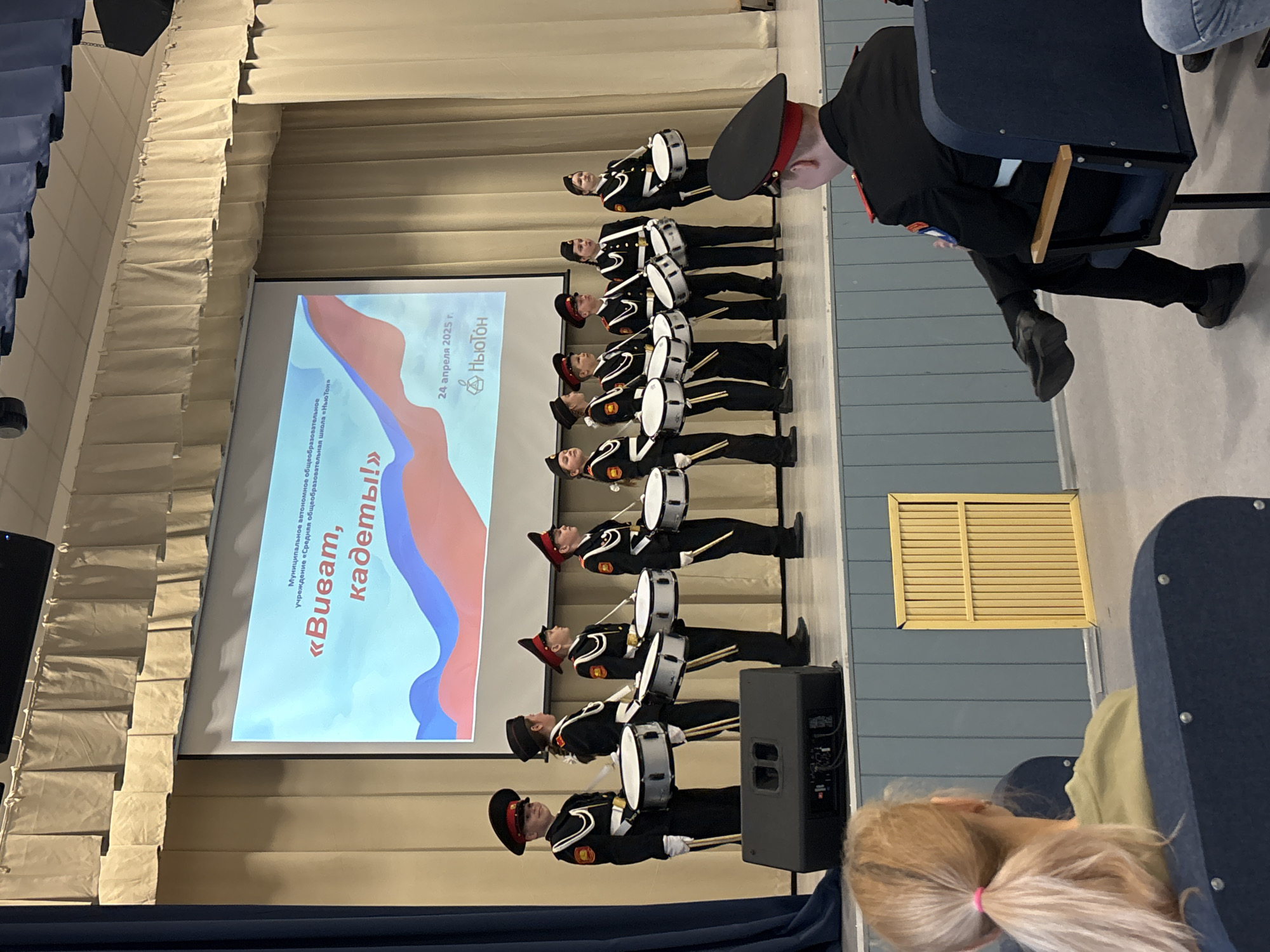Here is a detailed description of the image:
**Scene:** The image captures a performance or presentation taking place on a stage, likely inside an auditorium or similar venue.
**People:** There is a line of young individuals dressed in cadet uniforms, each holding and seemingly prepared to play a drum. Their uniforms include black jackets, matching pants, and hats with red accents. Seated among the audience are several indistinct individuals, including a child with blonde hair in a ponytail in the foreground. One person in the foreground audience on the right seems to be wearing a cadet uniform as well.
**Objects:** Key objects are the drums carried by each cadet, a screen displaying a presentation with text and graphic elements (including what appears to be the Russian flag), stage curtains, seating in the audience, and a speaker on the stage.
**Actions:** The cadets are standing in formation. The audience is seated.
**Interactions:** The cadets are performing for the audience.
**Environment:** The setting is an indoor venue, potentially a school auditorium. Based on the screen text, the event is located in Chaikovsky, Russia. The venue has a stage, a screen for presentations, and audience seating.
**Text:** The text on the screen reads: "Municipal Autonomous General Educational Institution 'Newton Middle School'" followed by the title "Vivat, Cadets!" and the date "April 24, 2025." A logo of a geometric shape and the word "Newton" are also visible on the screen.