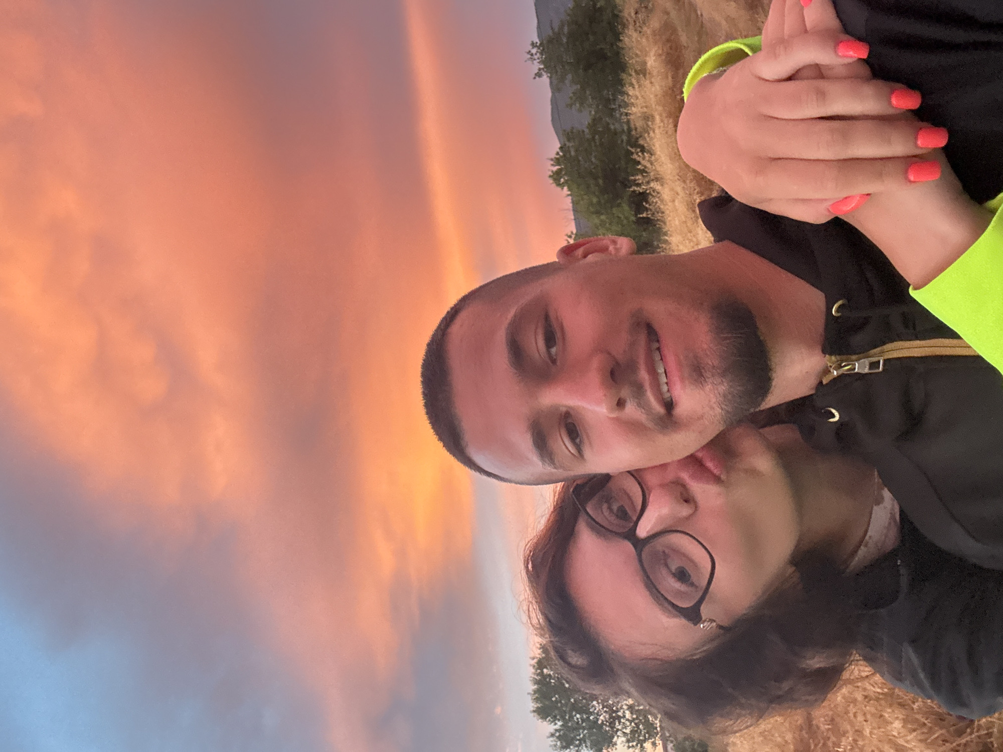A young couple is taking a selfie outdoors during what appears to be sunset. The sky is filled with vibrant hues of orange, pink, and purple, suggesting a beautiful evening. The background shows a grassy, dry field and some trees, with a hint of mountains in the distance.
The man in the foreground is smiling widely and looking at the camera. The woman is nestled beside him, with her head tilted and her eyes closed as if enjoying a tender moment. She is wearing glasses. Both are wearing casual clothing. The woman's nails are painted a bright coral color, and she is holding the man's hand. The overall mood of the image is romantic and joyful, capturing a moment of affection between the couple. There is no visible text in the image.