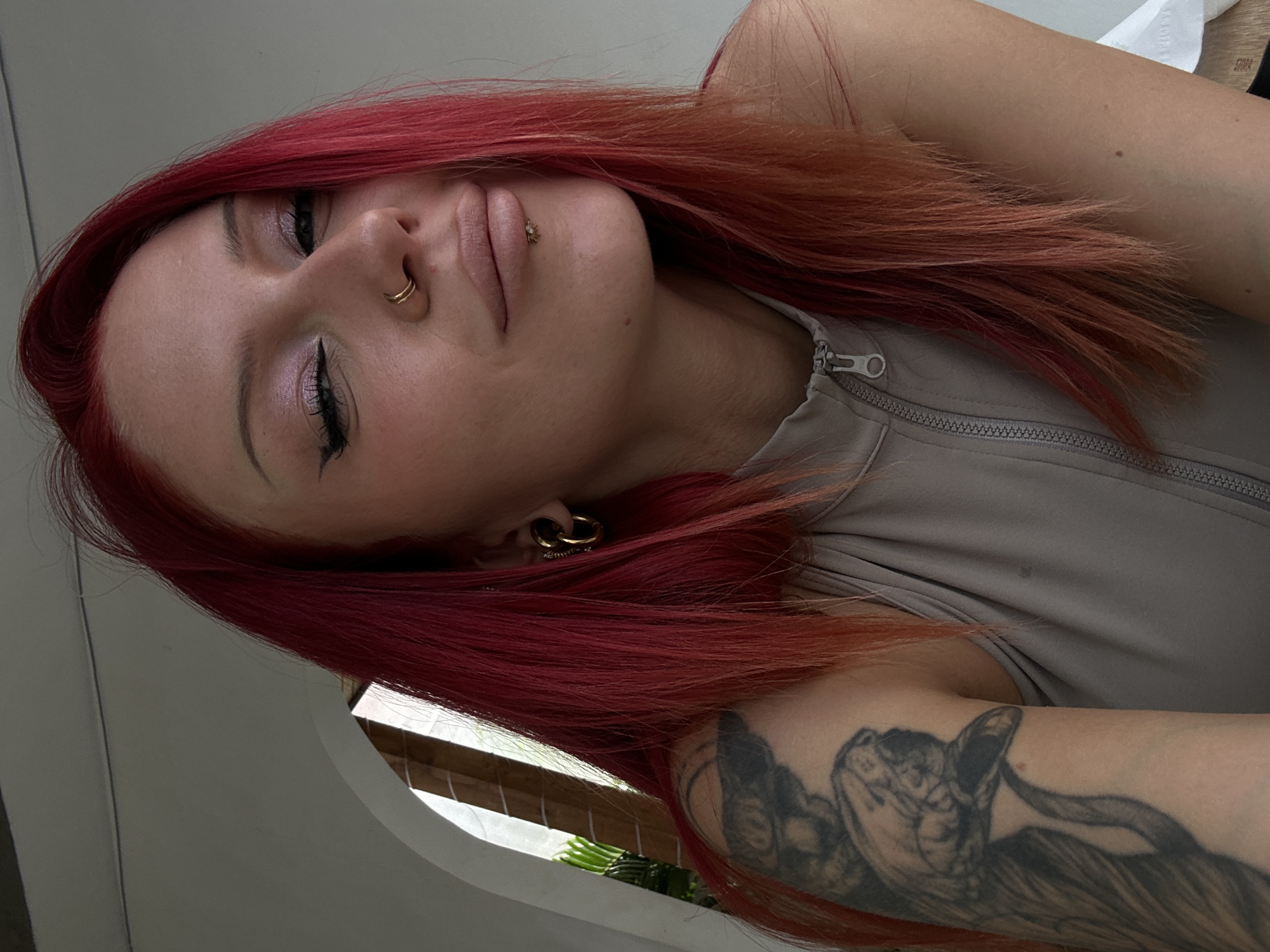 A woman with vibrant red hair is pictured in a close-up, head-and-shoulders shot. She is looking towards the camera with a slight smile and her eyes are partially closed. Her makeup includes winged eyeliner and shimmering eyeshadow. She has a double hoop nose ring and a piercing below her lower lip. She is wearing a grey, zippered top and has a large tattoo of a snake on her left arm. The background appears to be an indoor setting with a hint of an arched doorway and some greenery visible. The lighting suggests it is daytime.