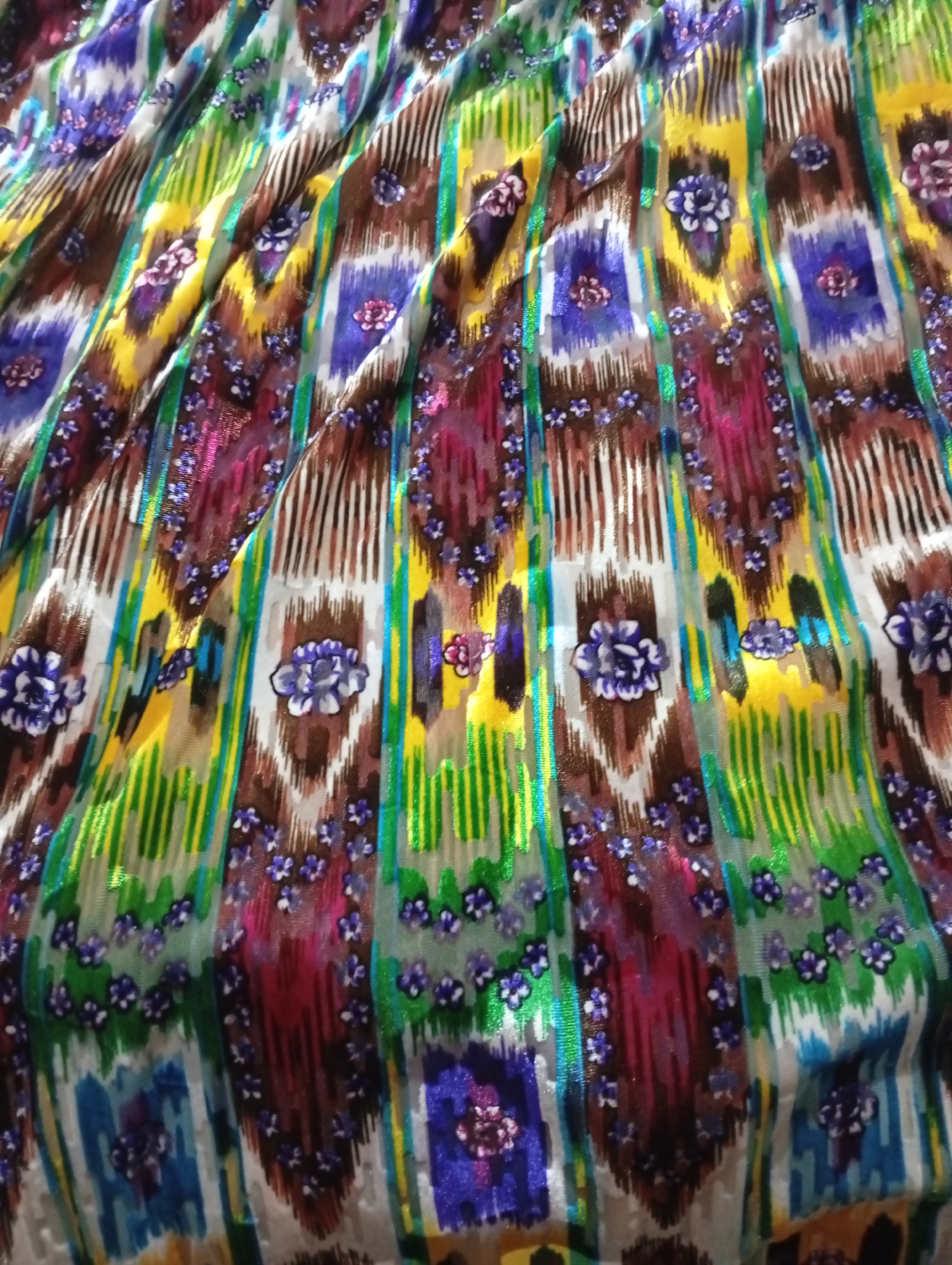 The image displays a close-up, overhead view of a colorful, patterned fabric. The fabric is draped, creating folds and shadows that highlight its texture and sheen. The pattern consists of vertical stripes of varying colors and widths, including shades of yellow, green, brown, purple, and blue. Within these stripes, there are smaller floral motifs, predominantly in shades of purple and white, and rectangular blocks of color. The fabric appears to be made of a synthetic material with a somewhat glossy finish, suggesting it could be satin or a similar material. There is no indication of a specific location within Seýdi, Turkmenistan; the image solely focuses on the fabric itself.