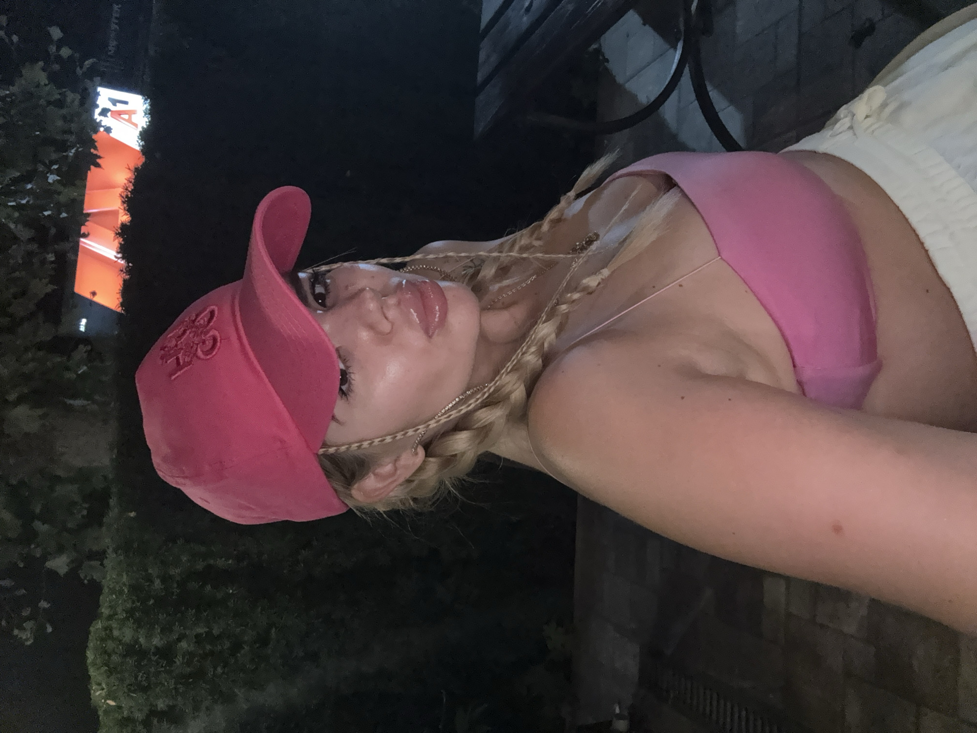 A woman with blonde hair braided into two pigtails, wearing a pink baseball cap and a pink tank top, poses for a selfie. She is outdoors at night. In the background, a lit sign with the letters "A1" is visible, along with the word "MONSTER" on a distant sign. There is foliage and a dark background suggesting it is a wooded or overgrown area. To the right, a dark wooden table and bench are partially visible. The woman has on white pants. Her lips are glossy, and she is wearing a necklace.