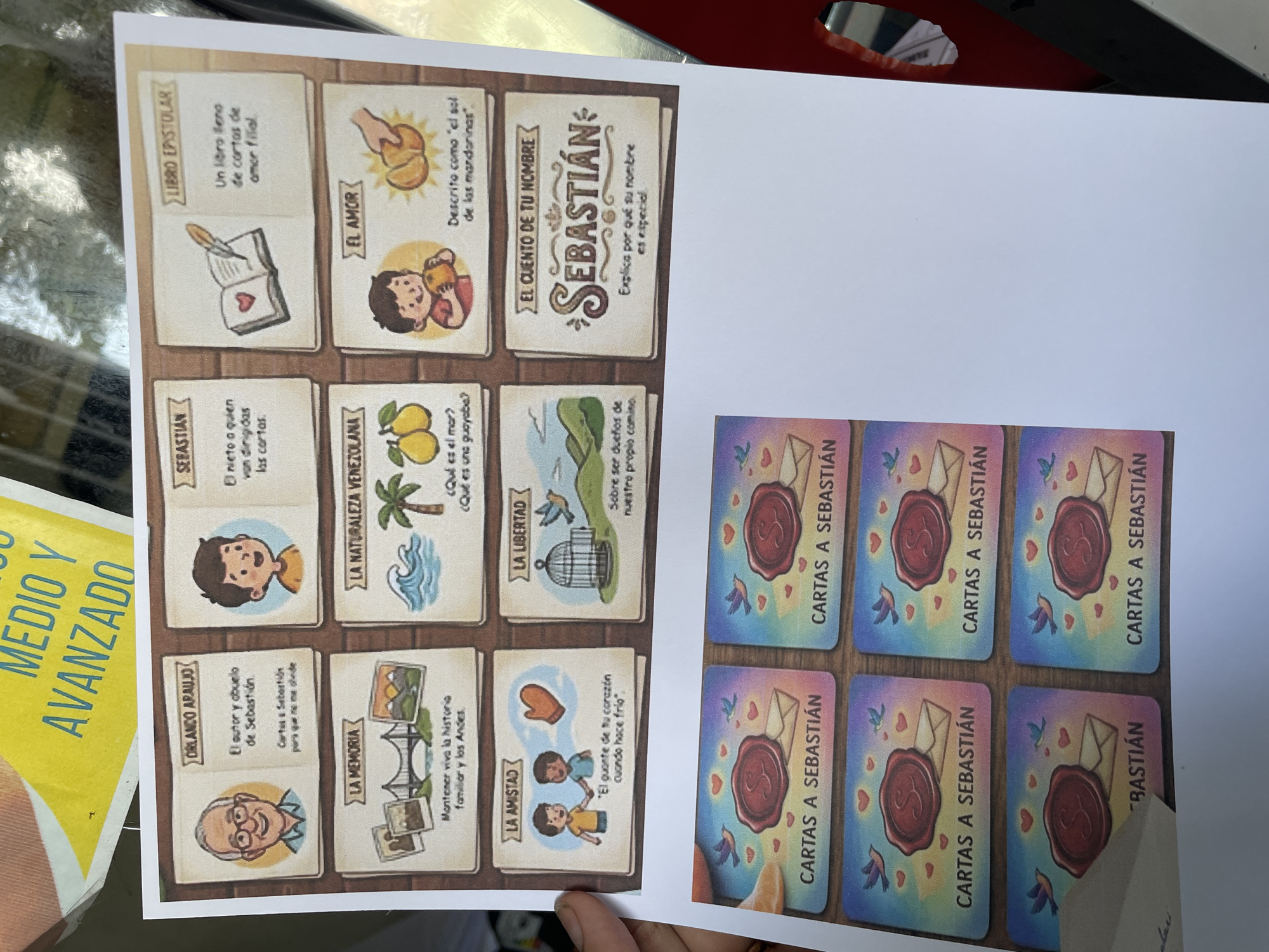 A printed sheet of paper, approximately A4 size, is captured in a slightly angled, downward-looking view. The sheet displays two distinct sections of graphics and text. The upper section features a 3x3 grid of nine illustrated squares, each containing a title, an image, and descriptive text in Spanish, generally relating to a book titled "Cartas a Sebastián" (Letters to Sebastián). 1. **Top Left:** Titled "ORLANDO ARAUJO," depicting a cartoon of an elderly man with glasses, described as "El autor y abuelo de Sebastián. Cartas a Sebastián para que no me olvide." (The author and grandfather of Sebastián. Letters to Sebastián so he doesn't forget me.) 2. **Top Middle:** Titled "SEBASTIÁN," depicting a cartoon of a young boy, described as "El nieto a quien van dirigidas las cartas." (The grandson to whom the letters are addressed.) 3. **Top Right:** Titled "LIBRO EPISTOLAR" (Epistolary Book), showing an open book with a heart and a feather quill pen, described as "Un libro lleno de cartas de amor filial." (A book full of letters of filial love.) 4. **Middle Left:** Titled "LA MEMORIA" (Memory), displaying multiple photographs including a bridge and a landscape, with the text "Mantener viva la historia familiar y los Andes." (To keep alive the family history and the Andes.) 5. **Middle Middle:** Titled "LA NATURALEZA VENEZOLANA" (Venezuelan Nature), showing an ocean wave, a palm tree, and two yellow fruits, with the questions "¿Qué es el mar? ¿Qué es una guayaba?" (What is the sea? What is a guava?) 6. **Middle Right:** Titled "EL AMOR" (Love), illustrating a boy holding a mandarin orange and a hand peeling another, described as "Descrito como 'el sol de las mandarinas'." (Described as 'the sun of the mandarins'.) 7. **Bottom Left:** Titled "LA AMISTAD" (Friendship), depicting two boys and a single mitten, with the text "'El guante de tu corazón cuando hace frío'." ('The mitten of your heart when it's cold'.) 8. **Bottom Middle:** Titled "LA LIBERTAD" (Liberty), showing an open birdcage with birds flying towards a mountainous landscape, described as "Sobre ser dueños de nuestro propio camino." (About being masters of our own path.) 9. **Bottom Right:** Titled "EL CUENTO DE TU NOMBRE" (The Story of Your Name), featuring a stylized "SEBASTIÁN" title, with the text "Explica por qué su nombre es especial." (Explains why his name is special.) The lower section of the sheet contains six identical small square graphics arranged in two