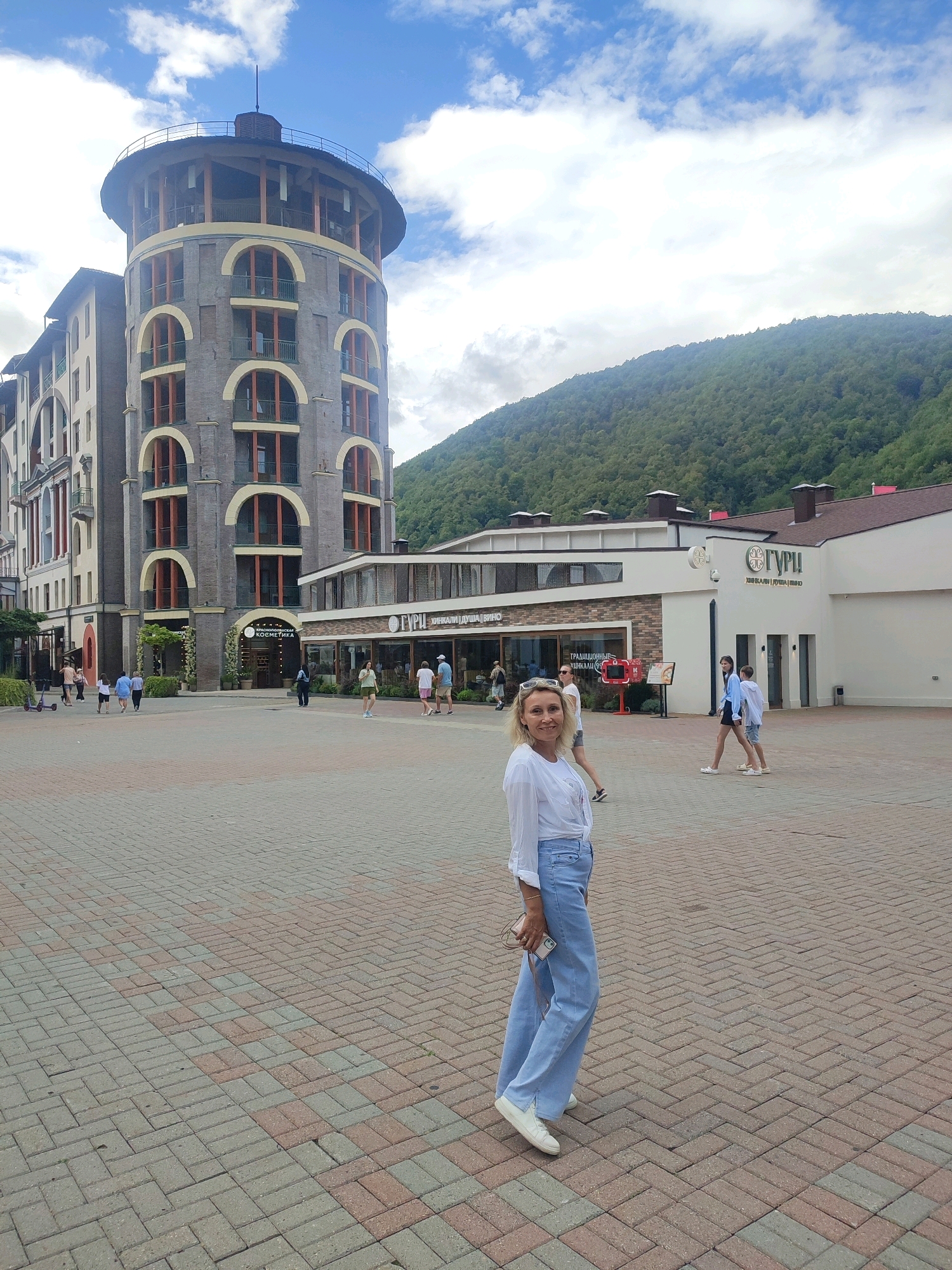 In the city of Estosadok, Russia, a blonde woman in a white shirt and light blue flared jeans is posing for a photo in a paved square. She is holding a small clutch bag in her left hand and her smartphone in her right. The square is made of brick pavers in shades of red and gray. Behind her, a circular building with arched windows and balconies stands tall, a characteristic architectural style of the area. Further in the background, a green, tree-covered mountain slopes upwards under a cloudy blue sky. To the right of the circular building, a modern white structure with a brown roof features the Cyrillic sign "ГУРЧИ" (GURCHI). Several people are walking in the background, suggesting a public gathering space.