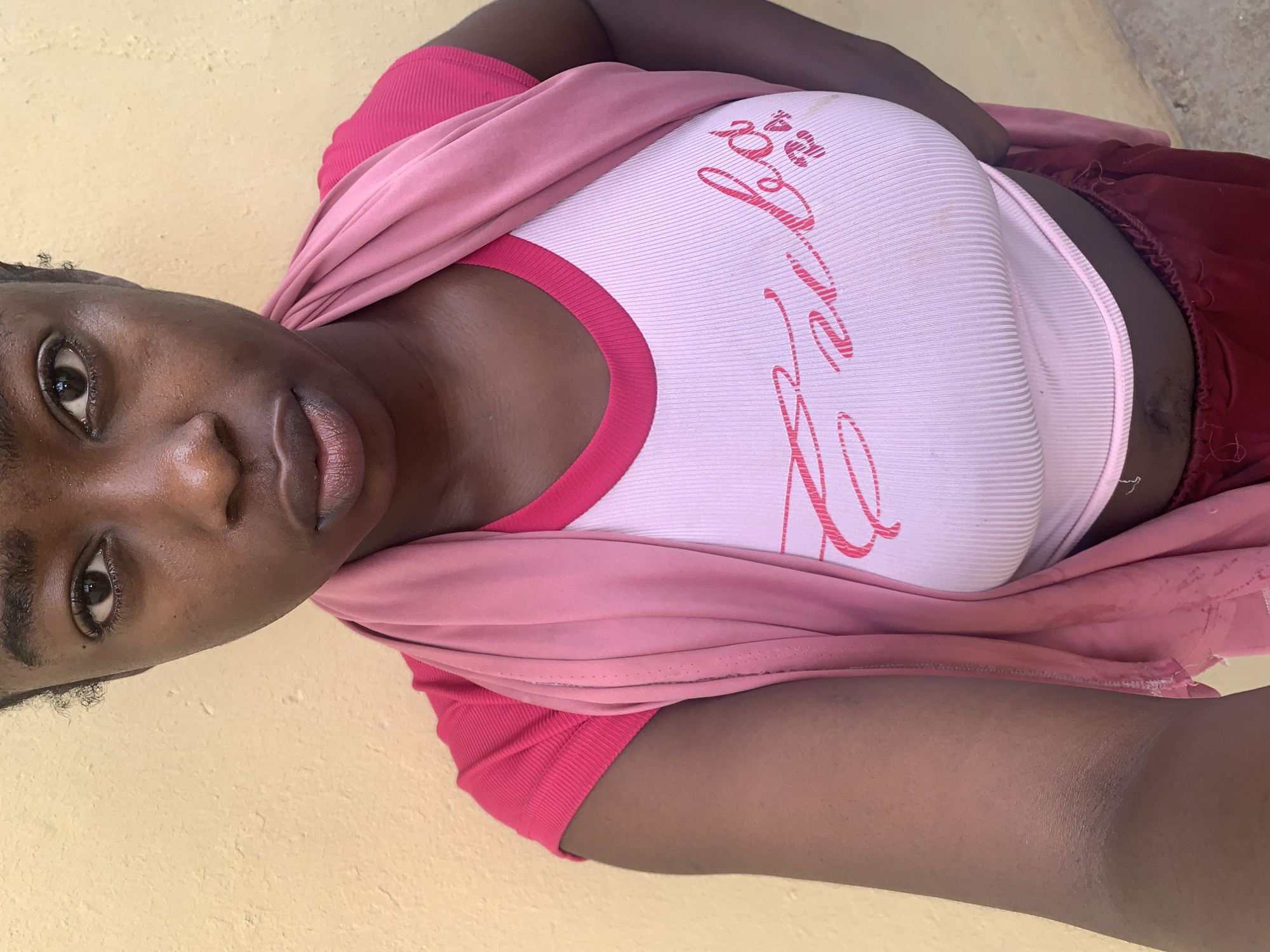 A medium-shot photograph of a Black African woman posing in an indoor setting, possibly in Rafin Par, Nigeria. The woman is wearing a pale pink ribbed tank top with a darker pink neckline and a script design on the front, layered under a darker pink t-shirt and a light pink scarf-like garment draped around her shoulders. She is wearing a reddish-brown garment on her right hip. She has dark skin and her face is in focus, with detailed features. The background is a light yellow or beige wall. The photo is taken from a slightly high angle.