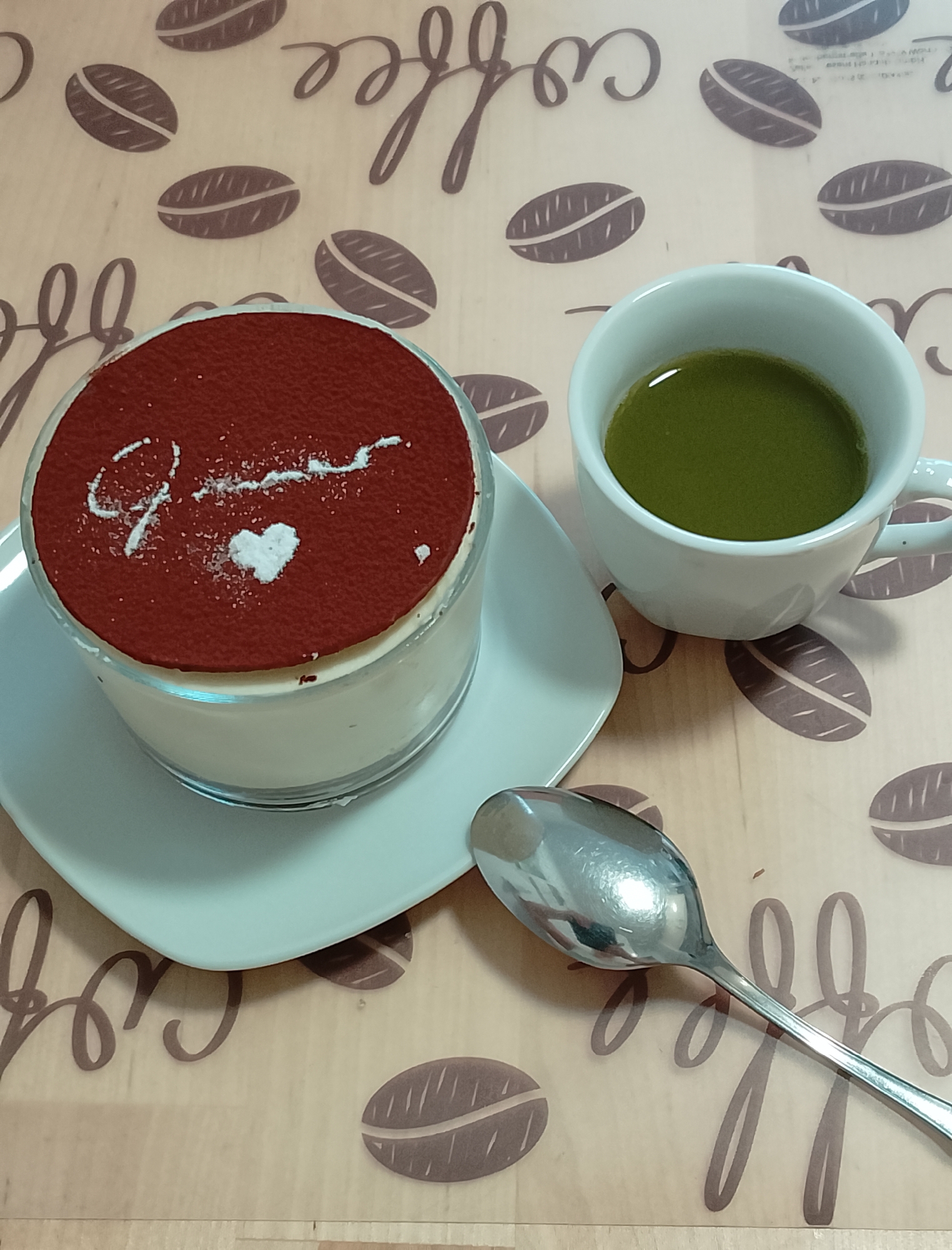 A close-up, overhead shot shows a dessert and a beverage on a table with a coffee bean-themed pattern. The dessert, in a glass dish, is topped with a dark powder, possibly cocoa, and features a white powdered heart and a swirled design. To the right of the dessert is a white ceramic mug filled with a vibrant green liquid, likely matcha tea. A silver spoon rests to the right of the dessert and its saucer. The background features a repeating pattern of coffee beans and the word "coffee" written in a script font.