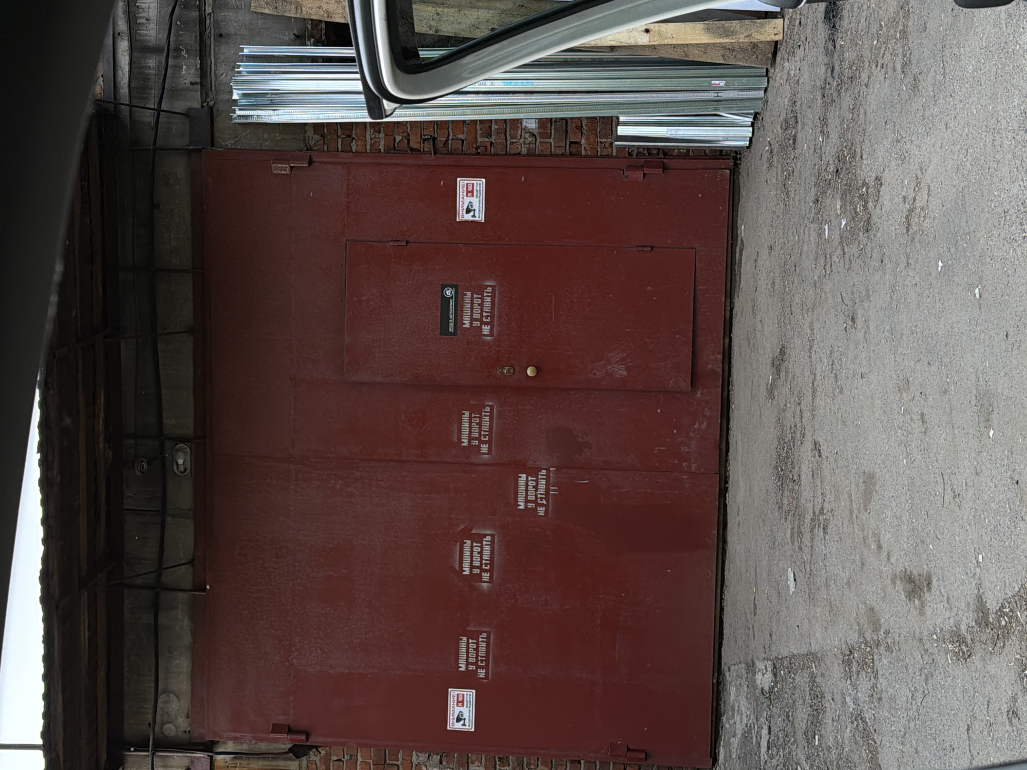 The image shows a red garage door with multiple signs in Russian that say "МАШИНЫ У ВОРОТ НЕ СТАВИТЬ," which translates to "Do not park cars in front of the gate." The door is set in a wall composed of exposed brick and deteriorating plaster. There is a stack of metal sheeting visible above the door, behind the brick. A dark, shadowed area with exposed wooden beams is on the left, and cracked concrete paving is on the right. Part of a vehicle is visible at the top of the image. The overall condition suggests an old or poorly maintained building. The location is listed as the city of Unknown, Unknown.