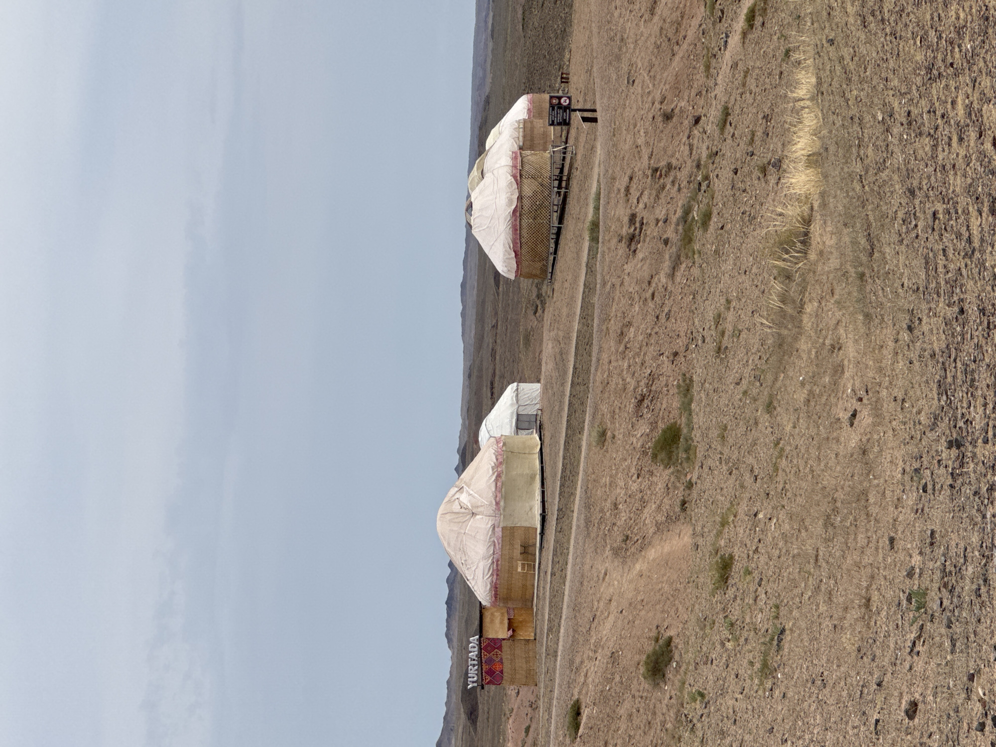 This is a medium-shot, eye-level, outdoor, daylight, full-color, vertical photograph of a desert landscape with three yurts. The yurts are traditional nomadic dwellings. The leftmost yurt is slightly larger and has "YURTADA" written in white text on a red and patterned panel. A small white tent is pitched behind it. Two other yurts are visible to the right, closer together and slightly further back. The landscape is arid and dry, with sparse brown vegetation and rocky soil. In the foreground, a dirt path winds up a small hill. The sky is overcast with light blue and grey clouds. The scene is located in Tamerlik, Kazakhstan.