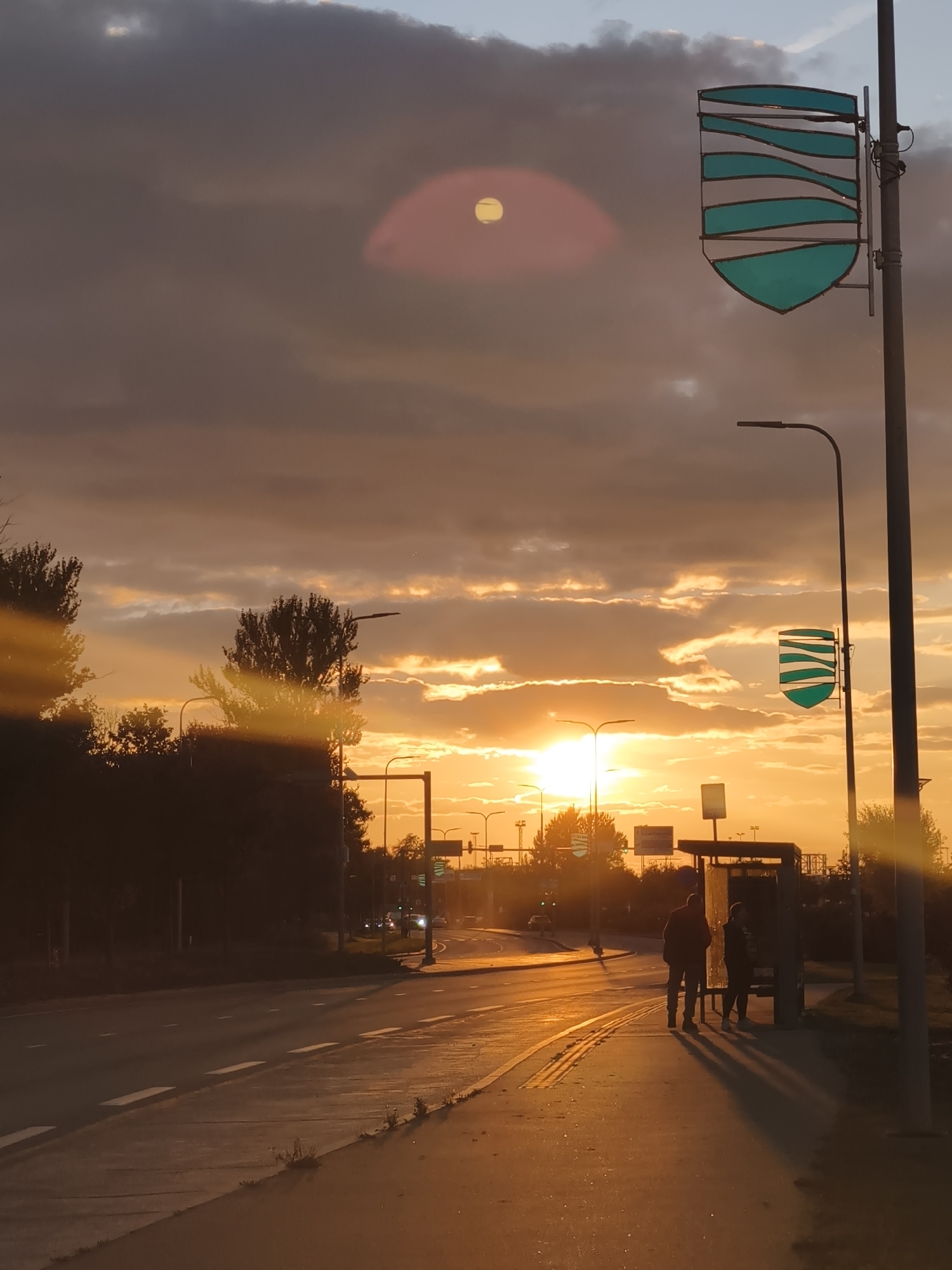 This is a photograph of a bus stop at sunset on a city street in Tallinn, Estonia. The sky is filled with dramatic clouds, with the sun casting a warm, golden glow over the scene. The sun is partially obscured by clouds, creating a lens flare effect. Two people stand at the bus stop, silhouetted against the bright sky. The bus stop itself has a modern design with a glass roof and side panels. A street lamp is visible to the right of the bus stop, and a street sign with a distinctive blue and white shield emblem is mounted on a pole. The road is wet, reflecting the light from the sunset, and the sidewalk is made of concrete. Trees line the street, and traffic lights and other street furniture are visible in the background. The overall impression is one of a serene and beautiful urban landscape at the end of the day.
