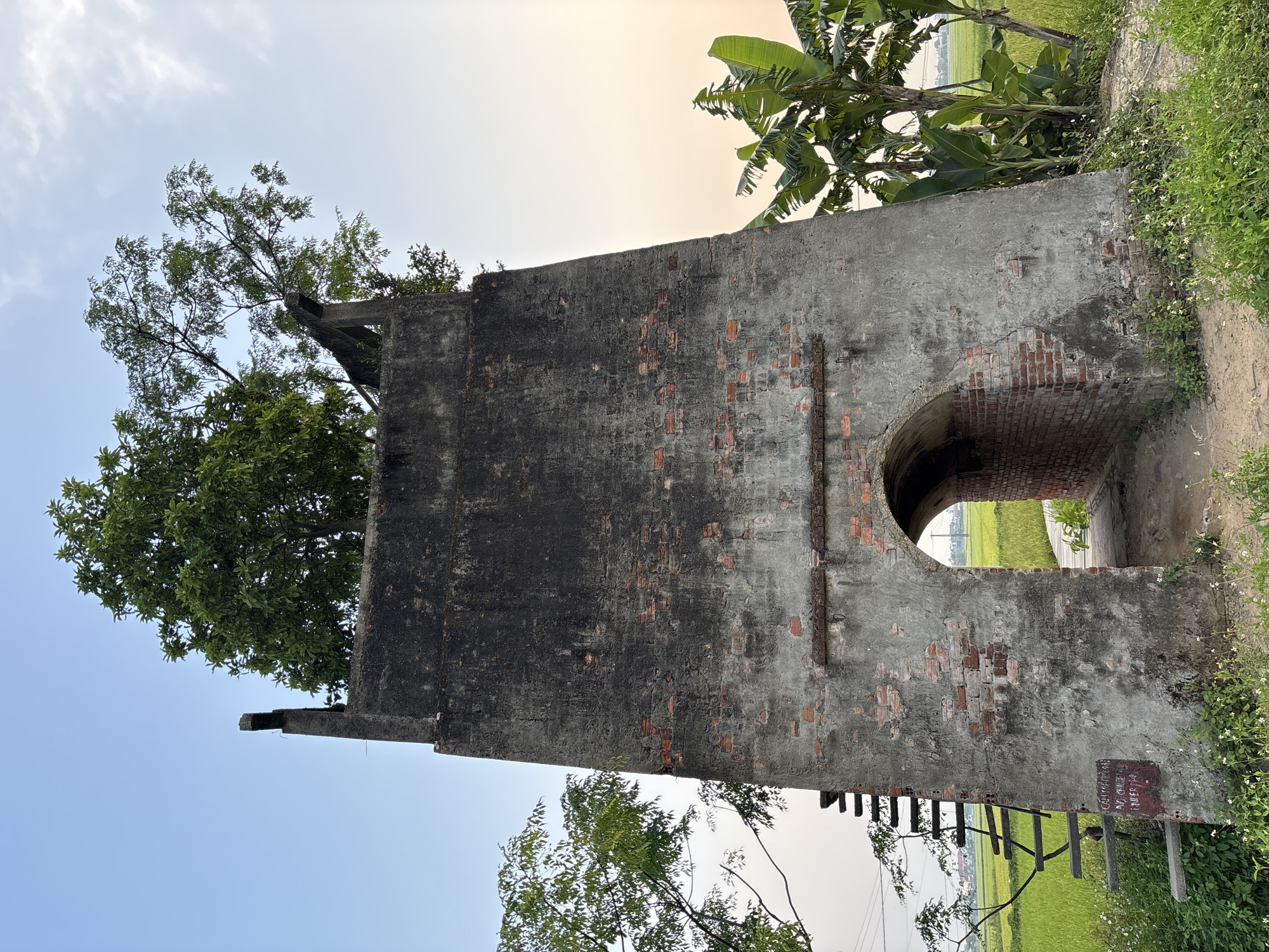 The image depicts a weathered, partially ruined brick structure with a large arched opening in a rural setting. Vegetation, including trees and grass, surrounds the structure. A ladder-like structure is leaning against the wall, and a small sign is visible near the base. The location is speculated to be in Đà Nẵng, Vietnam. The sky is overcast, with light clouds. The structure is the primary subject, and it appears to be an old fortification or similar historical building. No people are visible.