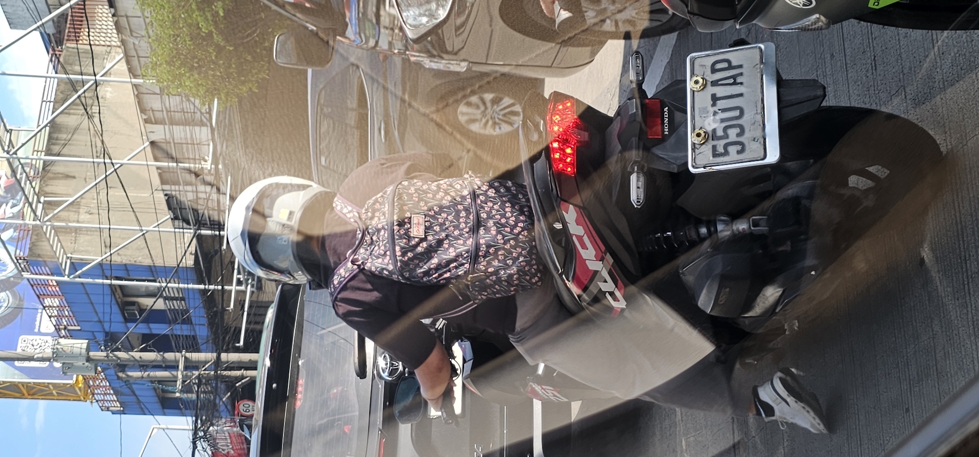 The image shows a motorcyclist from behind, wearing a black t-shirt and a patterned backpack, stopped in traffic in Quezon City, Philippines. The motorcycle has a license plate reading "550TAP". In the background, buildings and overhead wires suggest an urban environment.