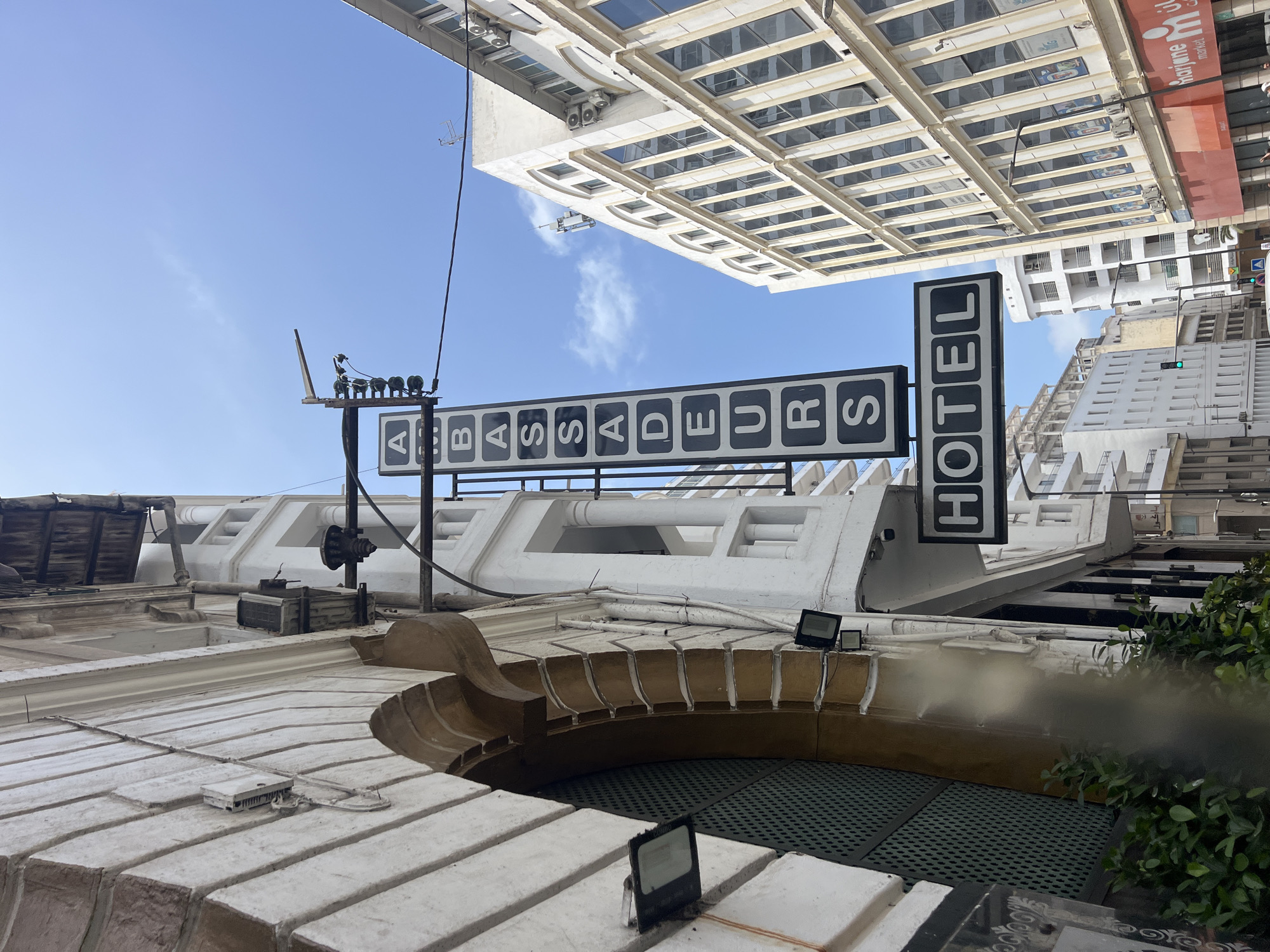 The image displays an upward-looking view of an urban environment, with the camera perspective rotated clockwise, causing buildings and the sky to appear tilted. In the midground, a large sign reads "HOTEL AMBASSADEURS," presented as a series of black rectangular panels with white uppercase letters. This sign is mounted on a white building facade, which features intricate architectural details, decorative cornices, and rooftop elements including exposed wiring, pipes, and spotlights. Green foliage is visible on the right side, extending from below the sign.
Above the hotel sign, a clear blue sky with a few scattered white clouds forms the background. Several modern, multi-story buildings rise into the sky, their facades featuring numerous windows. One distant building prominently displays a red and white sign with "Marjane Market" written in both English and Arabic. Other urban features such as traffic lights and external air conditioning units are also visible on the various structures.
The scene is brightly illuminated, indicating a clear, sunny daytime. There are no people visible, and no specific activities are taking place. The visible text includes "HOTEL AMBASSADEURS" and "Marjane Market." The architecture and clear skies suggest an urban setting in Casablanca, Morocco.