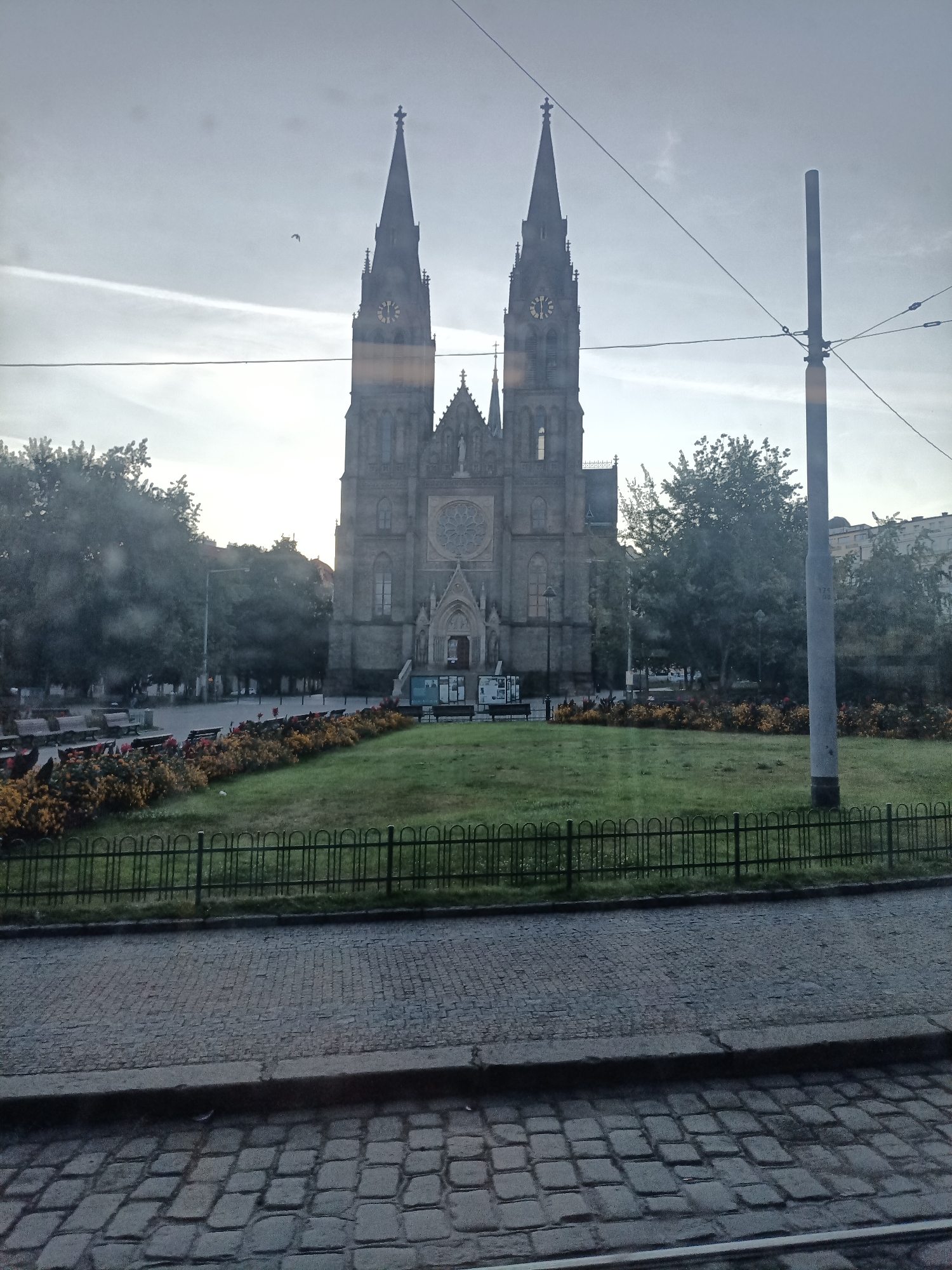 The image depicts the Basilica of St. Peter and St. Paul, located in Vyšehrad, Prague, Czechia. The grand Gothic Revival structure features two prominent spires, each topped with a cross. A large circular rose window is visible on the facade. The church is set against a pale sky with thin, wispy clouds. In the foreground, a well-maintained green lawn is bordered by a colorful flowerbed. A dark metal fence separates the lawn from a cobblestone street or pathway. To the right of the church, a tall, grey cylindrical pole stands, likely a lamppost or utility pole, with overhead wires crossing the frame. Trees are visible on either side of the church and in the background. The overall scene suggests a serene urban park setting adjacent to a significant historical landmark.