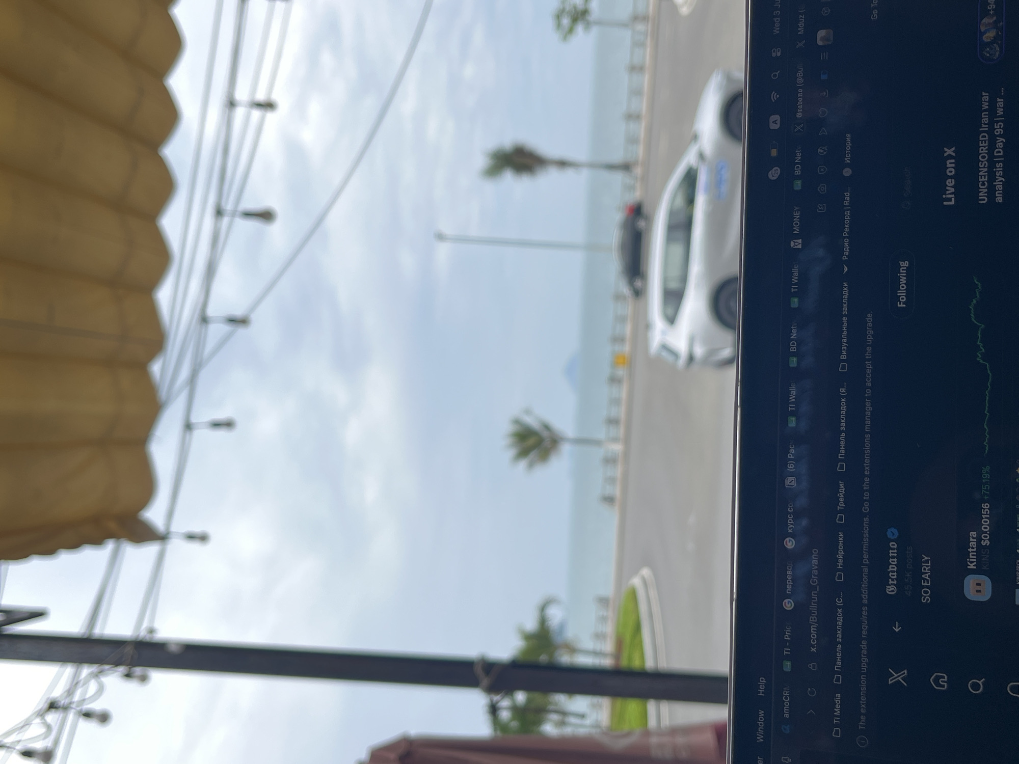 The image shows a digital screen, likely a laptop or tablet, displaying a web browser and social media content, positioned to reflect an outdoor scene. The screen is oriented vertically, causing the text to appear rotated.
In the reflection and seen beyond the screen, a coastal environment is visible under an overcast or cloudy sky. There's a wide body of water (presumably the sea or ocean) on the right, bordered by a road. Palm trees are scattered along the coastline, and a white car is prominent on the road, with another darker car partially visible behind it.
In the immediate foreground, part of a light brown or beige awning-like structure is visible on the left and top, with strings of lights or decorative cables hanging underneath. These elements suggest the device user is seated in a semi-outdoor or indoor area with an open view.
The digital screen displays a web browser with numerous tabs open, including "TI-Pric", "TI Media", "TI Wallet", "BD Netv", "MONEY", and several with Russian text like "Визуальные закладки" (Visual bookmarks), "Панель закладок" (Bookmarks panel), "Нейронки" (Neurons), "Трейдиг" (Trading), and "История" (History). An alert message reads, "The extension upgrade requires additional permissions. Go to the extensions manager to accept the upgrade." The social media platform X (formerly Twitter) is prominently displayed, showing a post by "@Grabano" with "45.5K posts" and the text "SO EARLY". Another section shows "Live on X" with "UNCENSORED Iran war analysis | Day 95 | war...". A financial ticker displays "Kintara KINS $0.00156 +75.19%". System information shows "Wed 3 Ju" (Wednesday, July) and "Window Help". The URL "x.com/Bullrun_Gravano" is also visible.
The scene combines a moment of digital engagement with a serene outdoor coastal backdrop, likely in a tropical or subtropical region like Bac Nha Trang Ward, Vietnam.