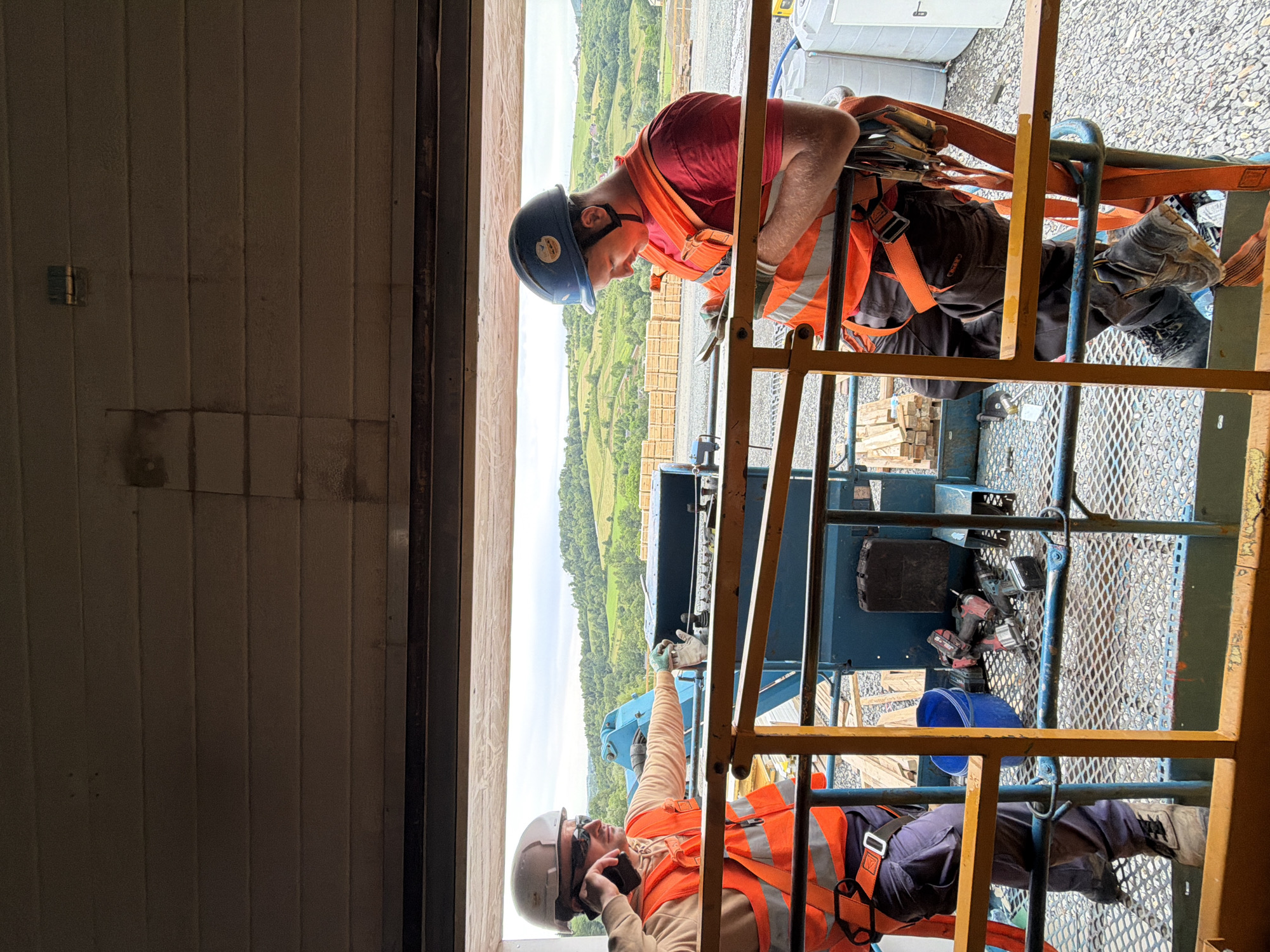 Two construction workers, both wearing orange high-visibility vests and hard hats, are on a raised platform overlooking a rural landscape with rolling hills. The worker on the left, wearing a white hard hat and a beige hoodie, is on a phone call while holding a pair of work gloves. The worker on the right, wearing a blue hard hat and a red shirt, is looking down at a mobile phone. Tools, including cordless drills, are visible on the platform between them. The background shows a construction site with stacked lumber and white containers, and beyond that, a verdant, hilly terrain suggesting the location is in the vicinity of Janowice, Poland. The overall scene depicts a moment of pause or communication during a construction project in an outdoor, semi-rural setting.