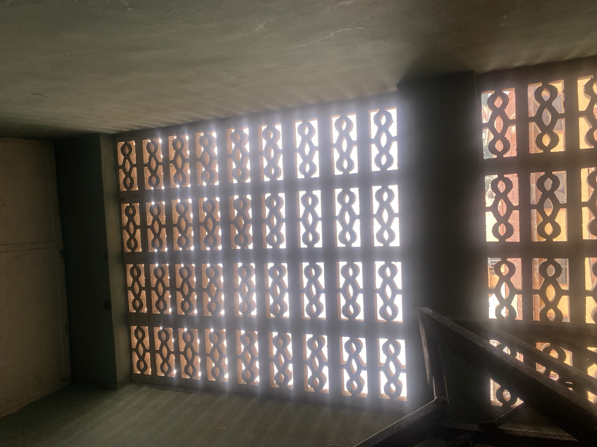 The image shows a view looking up through two levels of ornamental concrete block windows. The windows have a repeating pattern of interconnected loops. Sunlight streams through the openings, casting horizontal light streaks on the interior walls, which are painted a dull green. Part of a dark, angular metal structure, possibly a staircase railing, is visible in the lower left of the frame. Through the upper set of windows, the sky is visible, while the lower windows reveal the tops of buildings with reddish-brown tile roofs. The architectural style suggests an older building, possibly in a residential area. The scene appears to be located in the city of Abeokuta, Nigeria.