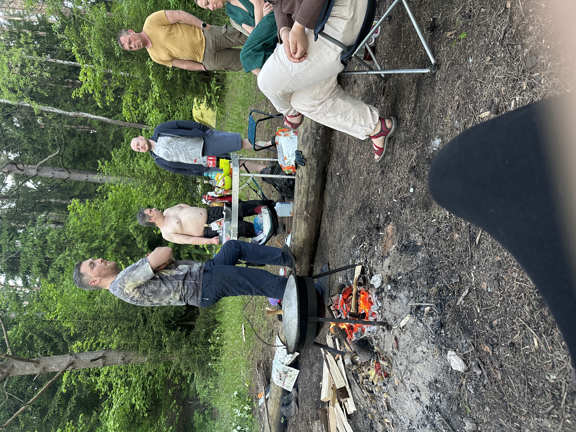The image depicts a group of people gathered outdoors, likely in a rural or wooded area near Юзуфоўскі сельскі Савет, Belarus. A fire is burning beneath a large cooking pot, attended by a man in a camouflage shirt and dark pants with his arms crossed. Several other men are present, some standing and one shirtless. A woman is seated on a folding chair. A table laden with drinks and food is visible in the background. The ground is covered in dirt, leaves, and wood debris. The scene suggests an informal gathering or cookout in a natural setting.