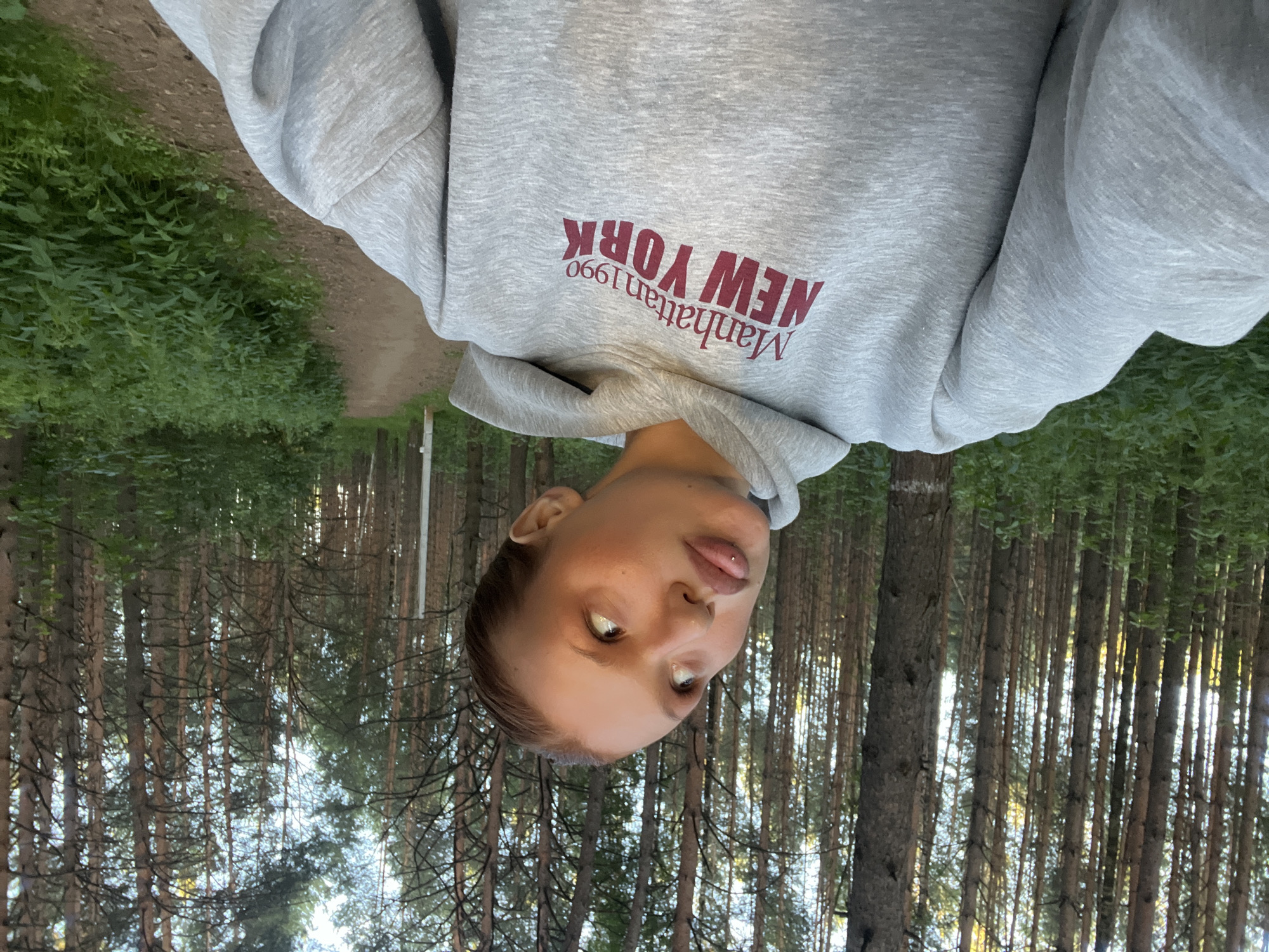 A young Caucasian woman with blonde hair pulled back is captured in a selfie taken in a forest. She is wearing a light grey hooded sweatshirt with the text "Manhattan 1990 NEW YORK" printed in a dark red font on the front. Her head is tilted back, and her eyes are looking upwards. Her lips are slightly parted, revealing a piercing on her lower lip. The background consists of a dense forest with tall, slender trees and lush green undergrowth. A dirt path is visible to the right of the woman. The lighting suggests it is either early morning or late afternoon. The scene appears to be located within the city of Chaikovsky, Russia, specifically in a forested area accessible by a path.