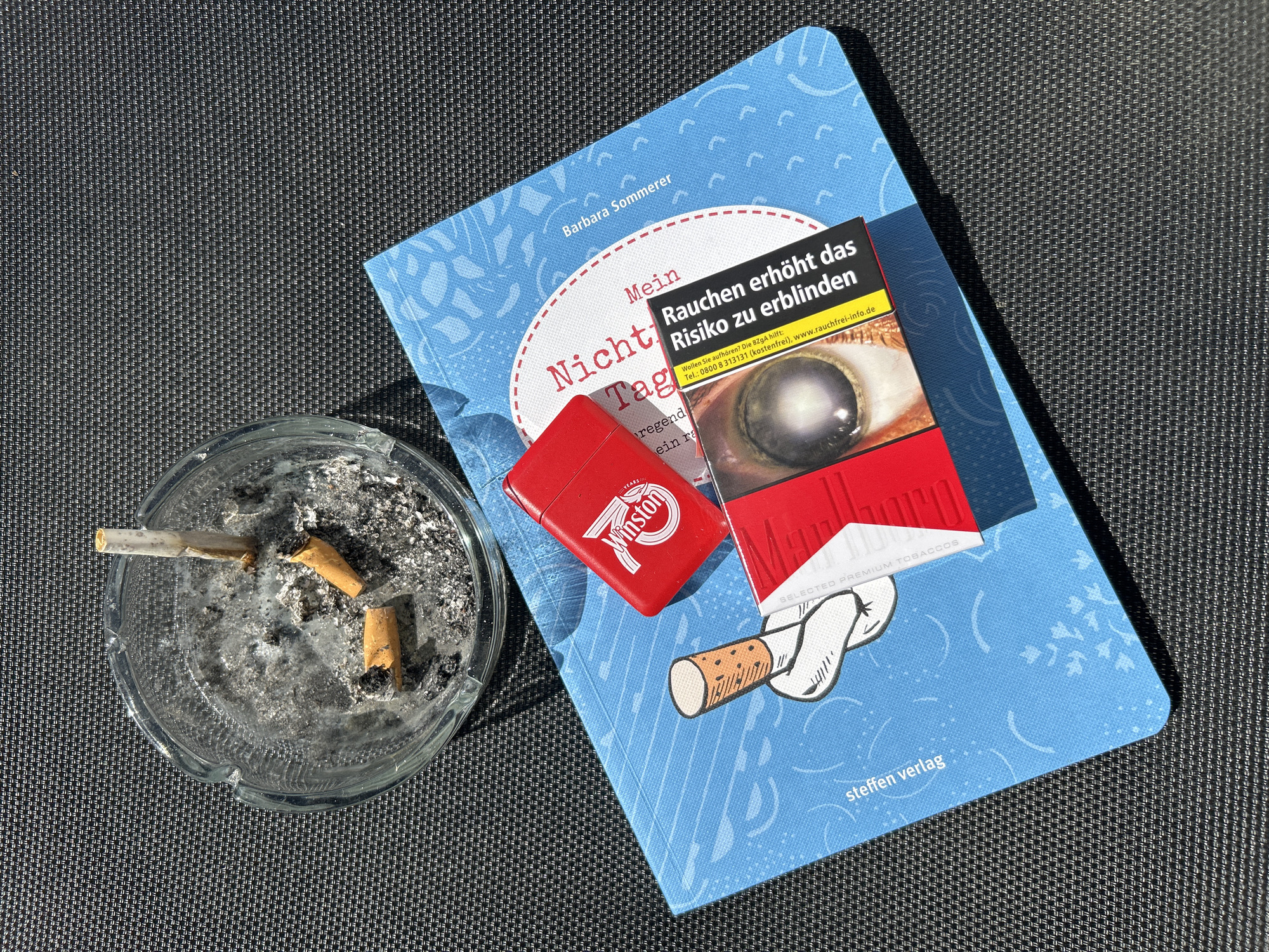 On a dark gray textured surface, a blue book titled "Mein Nicht-Tag" by Barbara Sommerer lies next to a clear ashtray filled with cigarette butts and ash. A red Winston lighter sits atop the book. A pack of Marlboro cigarettes with a health warning label depicting an eye disease is also placed on the book. The overall scene suggests smoking and reading.