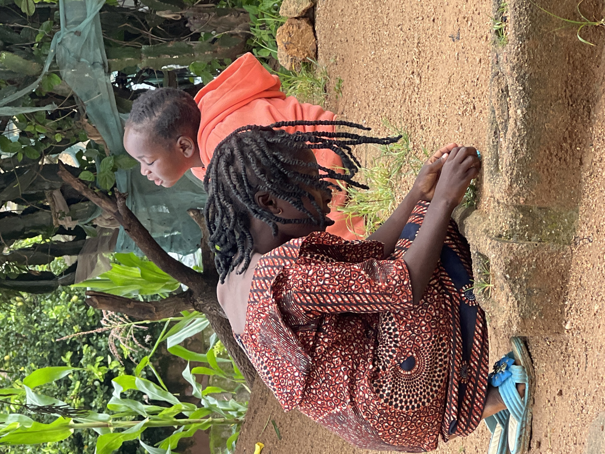 The image depicts two young individuals in an outdoor setting, likely a residential area or yard, within Jos, Nigeria. In the foreground, a person is kneeling, facing downwards with their head bowed, wearing a red and black patterned garment and blue sandals. Their hands are engaged with something on the ground. Behind them and to the right, another young person is visible from the chest up, wearing an orange hooded garment and looking upwards. The environment includes sandy ground, a stone border, various plants including corn stalks, and tree branches, with some green netting draped in the background. The lighting suggests it is daytime.