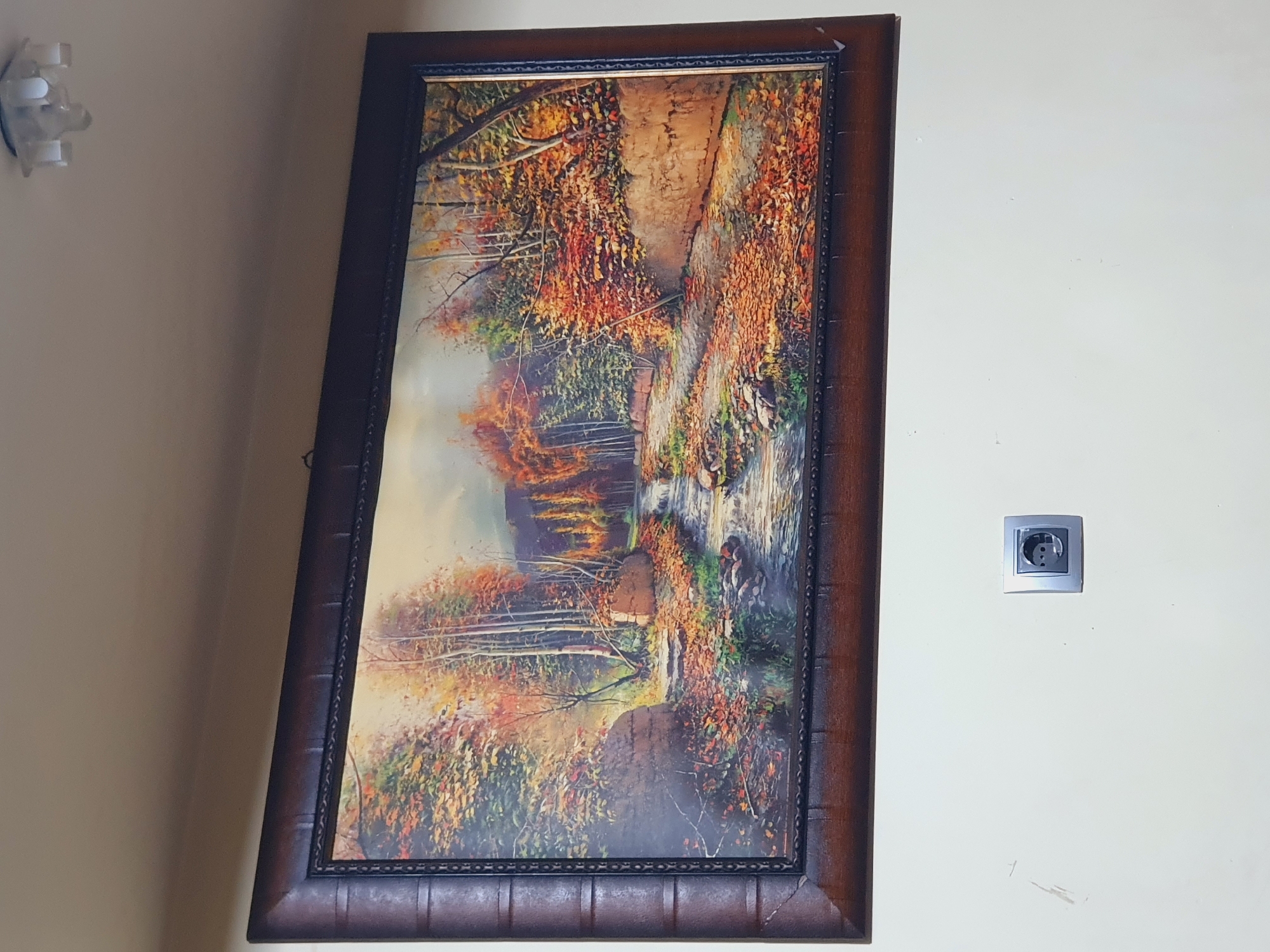 The image shows a framed painting of an autumn forest scene hanging on a light-colored wall. The painting depicts a stream flowing through a path covered with fallen leaves, surrounded by trees with colorful foliage. There is a small, silver electrical outlet mounted on the wall below the painting. The ceiling above the painting has a light fixture. The setting appears to be indoors, likely a residential or commercial space. No specific location in Shiraz, Iran, can be identified from the image.