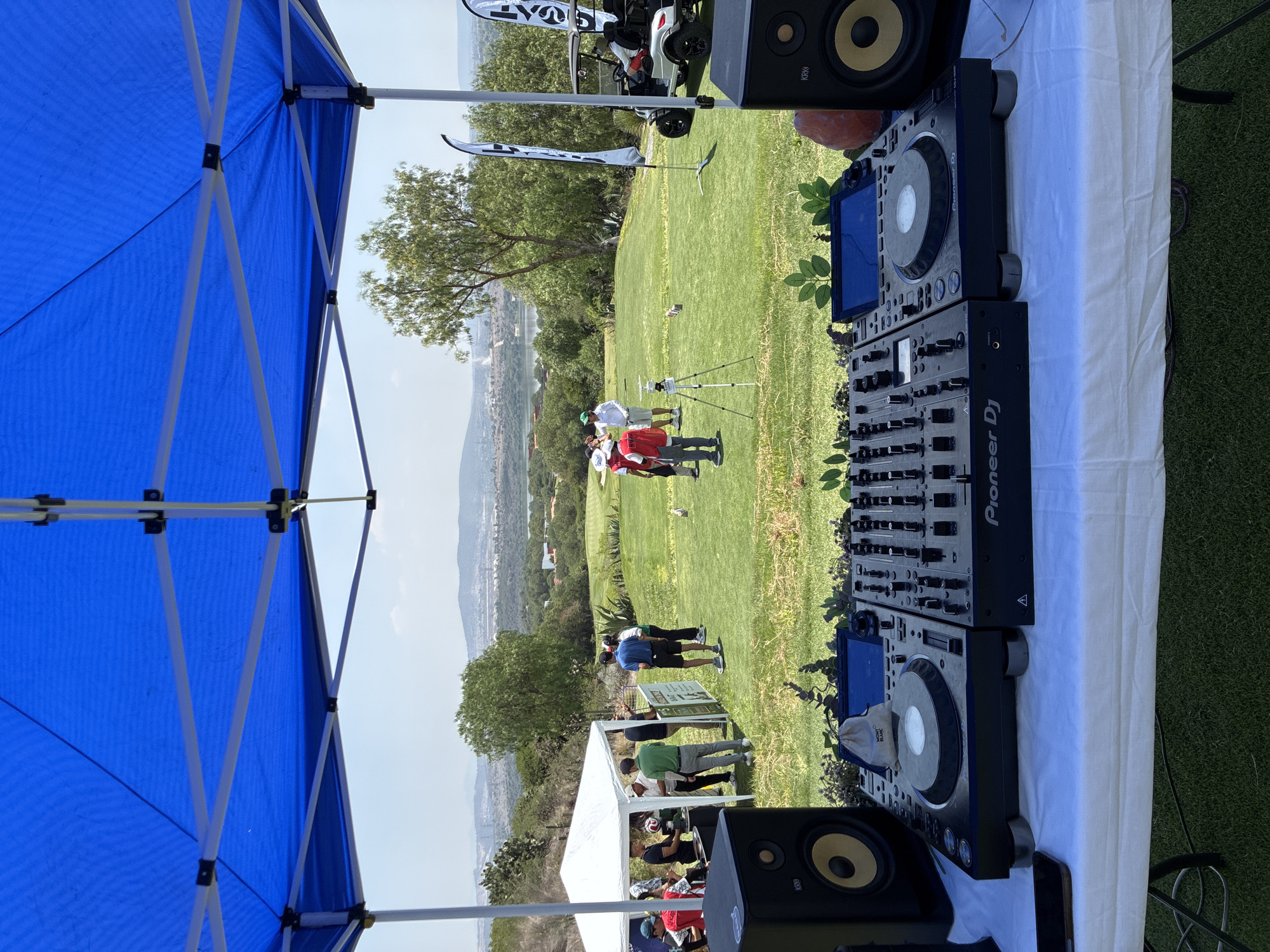The image depicts an outdoor event in Amanali, Mexico, likely a golf tournament or gathering, with a DJ setup in the foreground. The DJ equipment consists of two Pioneer CDJ units and a Pioneer DJ mixer placed on a white-covered table, flanked by black KRK speakers. A blue tent provides shade over the DJ area.
In the background, a green golf course extends towards a distant cityscape. Several groups of people are visible on the course, some wearing golf attire and others gathered around a white tent. A golf cart is parked near the top of the frame. The weather appears sunny and clear.