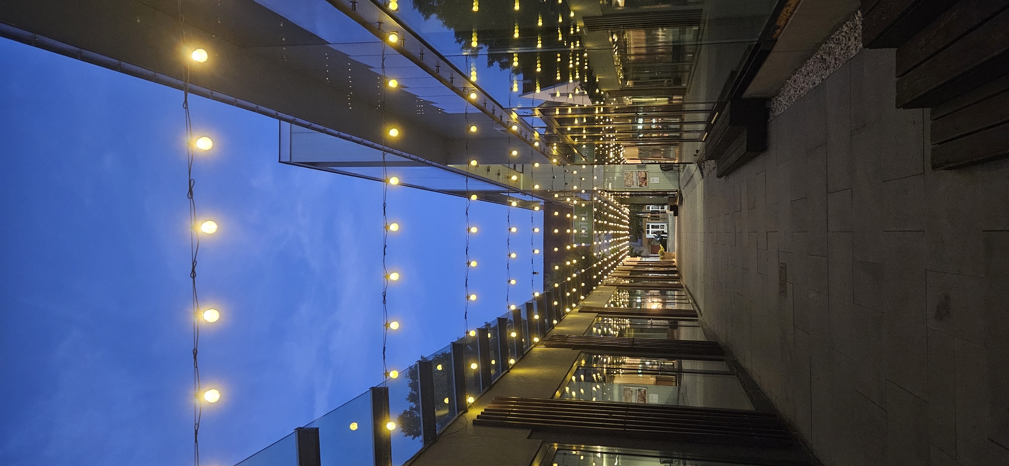 The image depicts an outdoor pedestrian walkway in Hévíz, Hungary, at dusk or dawn. The walkway is flanked by buildings with large glass facades, reflecting the evening sky and rows of string lights. These lights, composed of numerous round, warm-toned bulbs, are strung in parallel lines overhead, illuminating the path. The ground is paved with large, flat rectangular stones. Wooden benches are visible along the right side of the walkway. In the distance, a sign reading "HOTEL" is illuminated green. A few individuals are walking away from the camera in the background, suggesting a public or commercial area. The overall atmosphere is serene and inviting, with the artificial lighting creating a warm ambiance against the cool blue of the sky.
