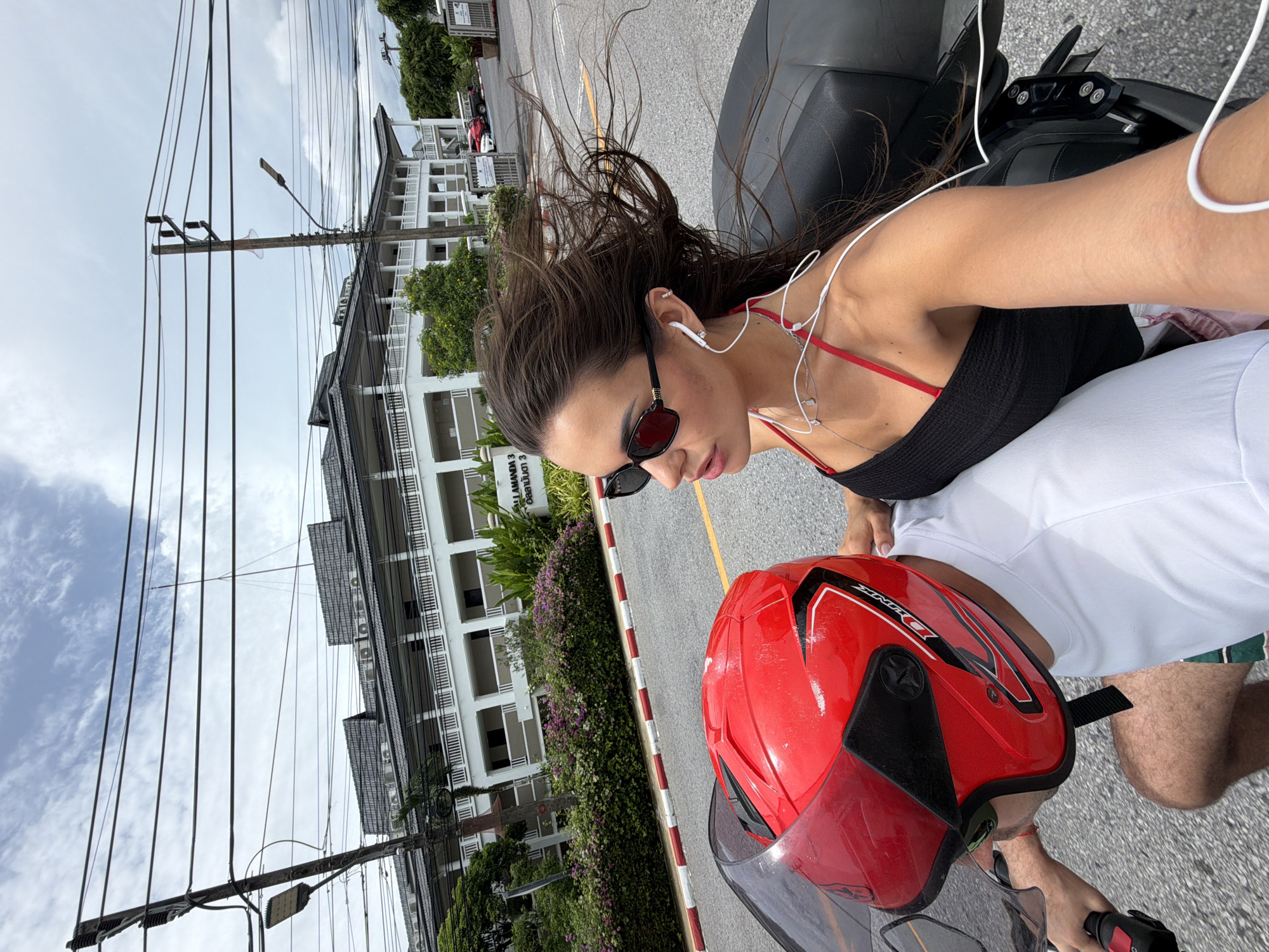 A woman with long brown hair, wearing sunglasses and a black top, is riding on a black motorcycle in Choeng Thale, Thailand. She has white headphones in her ears. In front of her, a person wearing a red helmet is driving the motorcycle. The road is lined with a white building with multiple balconies and green foliage. Power lines are visible above.