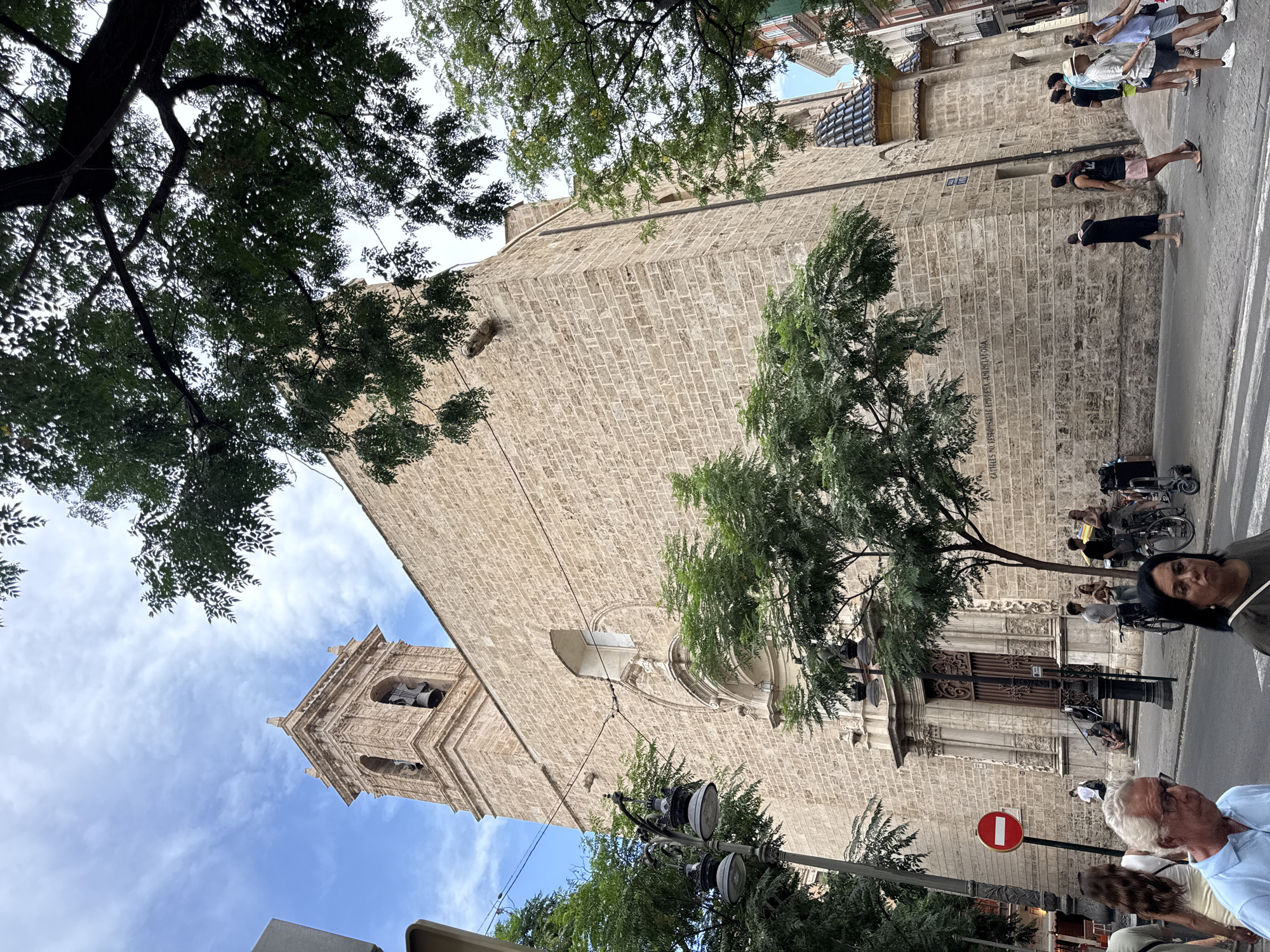 The image shows a street scene in Valencia, Spain, with a large stone church as the prominent background element. The church features a bell tower with an arched belfry containing two bells. The facade is constructed of light-colored stone blocks and has a large arched entrance with intricate wooden doors. Above the entrance, there's a round window and ornate architectural details. The church's roof has distinctive blue tiles.
In the foreground, people are walking on the sidewalk and street. Some are carrying shopping bags, others are talking, and a few are with bicycles. A street lamp with multiple globes stands to the left of the church. A red and white circular "no entry" sign is also visible on the left side of the street. Trees with lush green foliage partially obscure the view of the church, casting shadows on the building and the street. The sky is blue with scattered clouds. The scene suggests a normal day in a European city, with pedestrians and architectural landmarks.