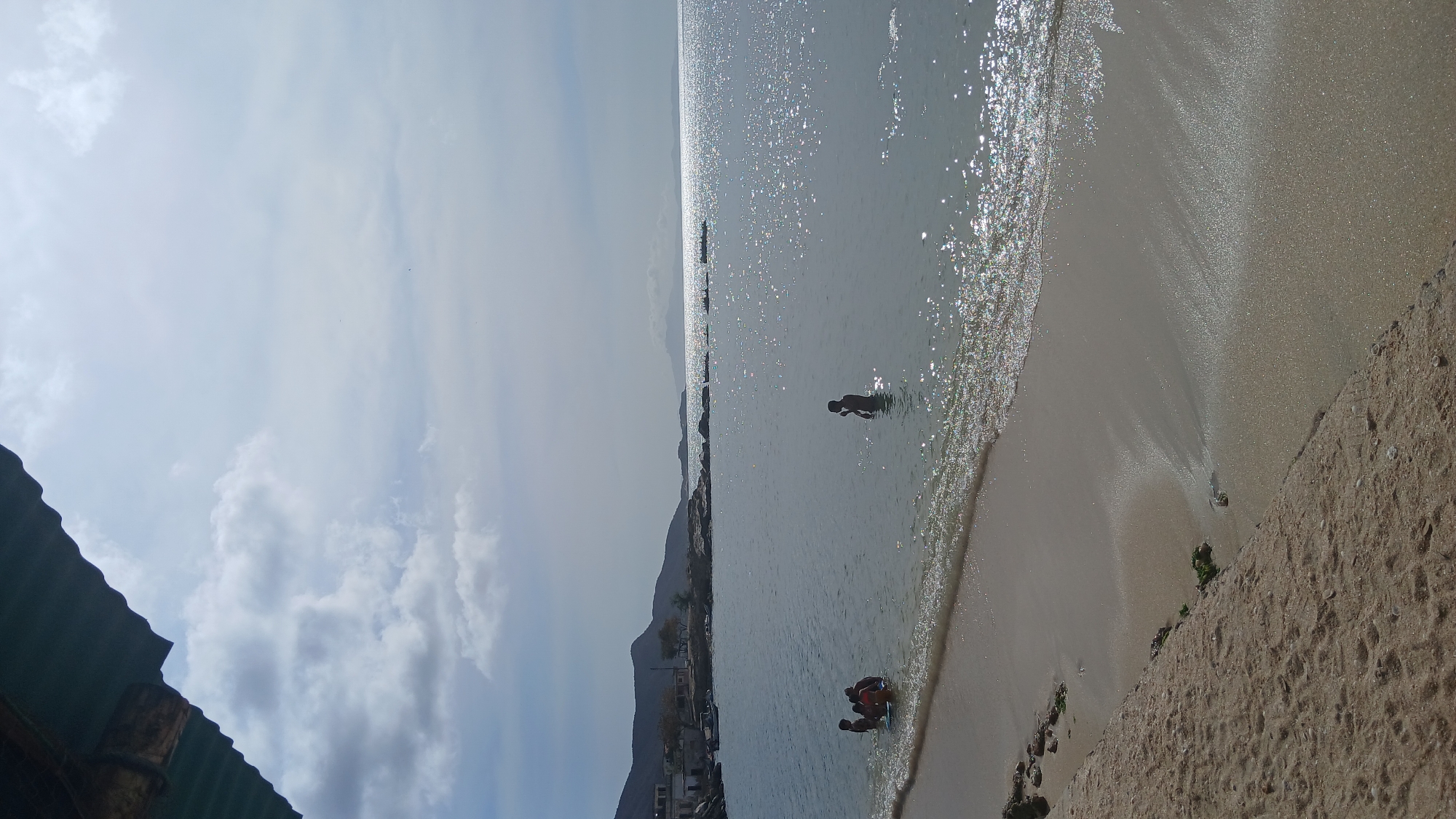 The image depicts a beach scene in Juan Griego, Venezuela. The focus is on the shoreline where the water meets the sand. The water is relatively calm with sunlight reflecting off its surface. A person stands alone in the water near the middle of the image. A group of individuals are clustered near the shore closer to the bottom of the image, some of whom appear to be children or young adults. The beach itself appears to be made of light-colored sand. In the distance, mountains and buildings are visible along the coast. The sky is partly cloudy. A section of a corrugated metal roof is visible in the lower left corner of the image.