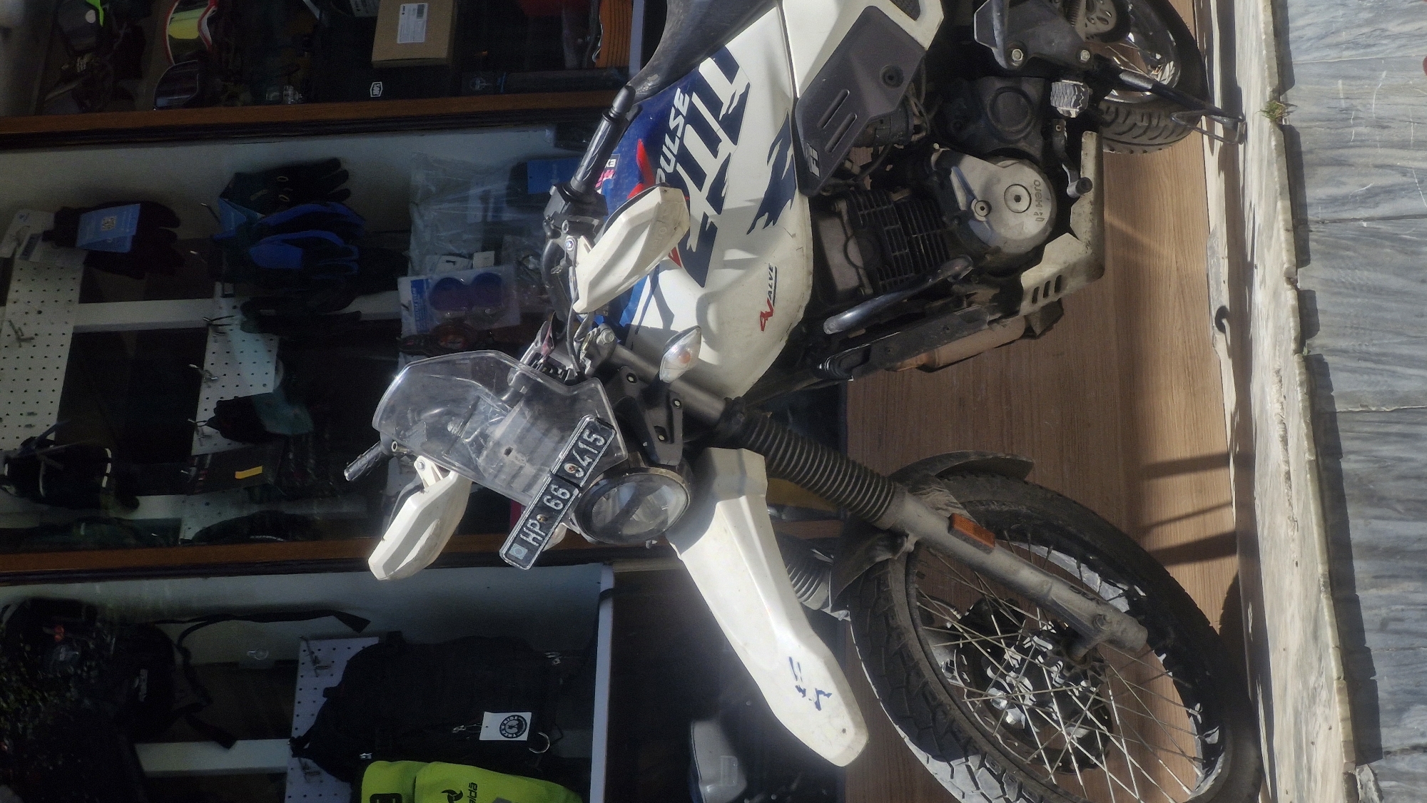 A white motorcycle is parked in front of a shop in Vashisht, India. The motorcycle has a license plate that reads "HP 66 9415." The shop appears to sell motorcycle gear, as evidenced by the helmets, gloves, and jackets visible on display inside. The motorcycle is a dual-sport model, suitable for both on-road and off-road riding. It has a tall windscreen, handguards, and knobby tires. The motorcycle is clean and in good condition. The shop is well-lit and organized. The environment appears to be a busy market area, with other shops and people visible in the background.