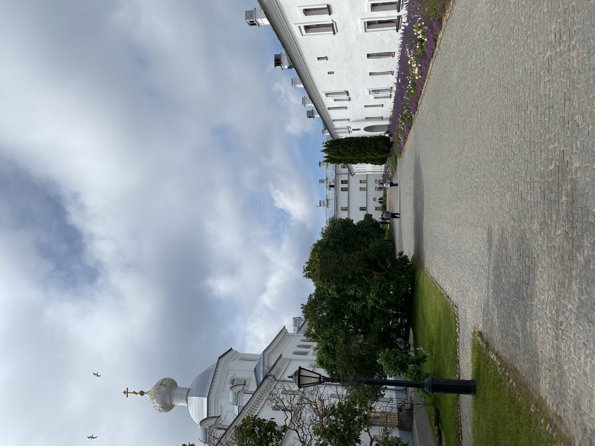 The image shows a view of the Valaam Monastery complex in Sortavala, Russia. In the foreground, a cobblestone pathway leads away from the viewer, bordered by green grass and mature trees on the left, and a white building with numerous windows on the right. Along the right side of the pathway, a flowerbed with purple and white flowers runs adjacent to the white building.
On the left side of the pathway, a white church with a golden dome topped with a cross is visible, along with a black lamppost. Further down the pathway, several people can be seen walking, and more white buildings characteristic of the monastery complex are visible in the distance. The sky above is cloudy with patches of blue sky. The overall scene depicts a serene and well-maintained courtyard within a historical religious site.