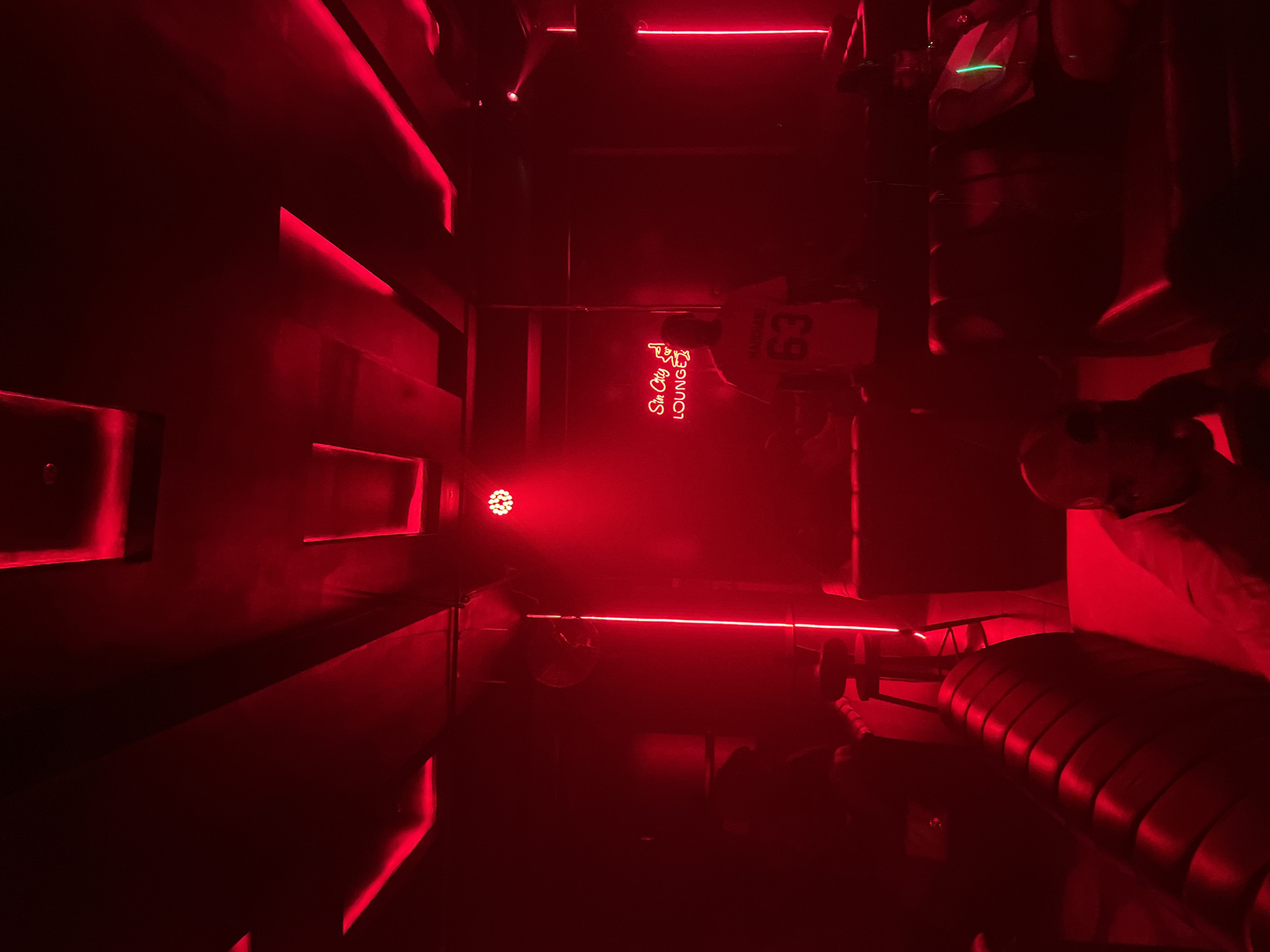The image depicts the interior of Sin City Lounge in Abuja, Nigeria. The room is illuminated with red light, highlighting architectural features such as layered wall panels and linear lights. A neon sign with "Sin City Lounge" is visible. Occupants, including people wearing jerseys with the number 63, are present. Furniture, like a padded bench, is also present.