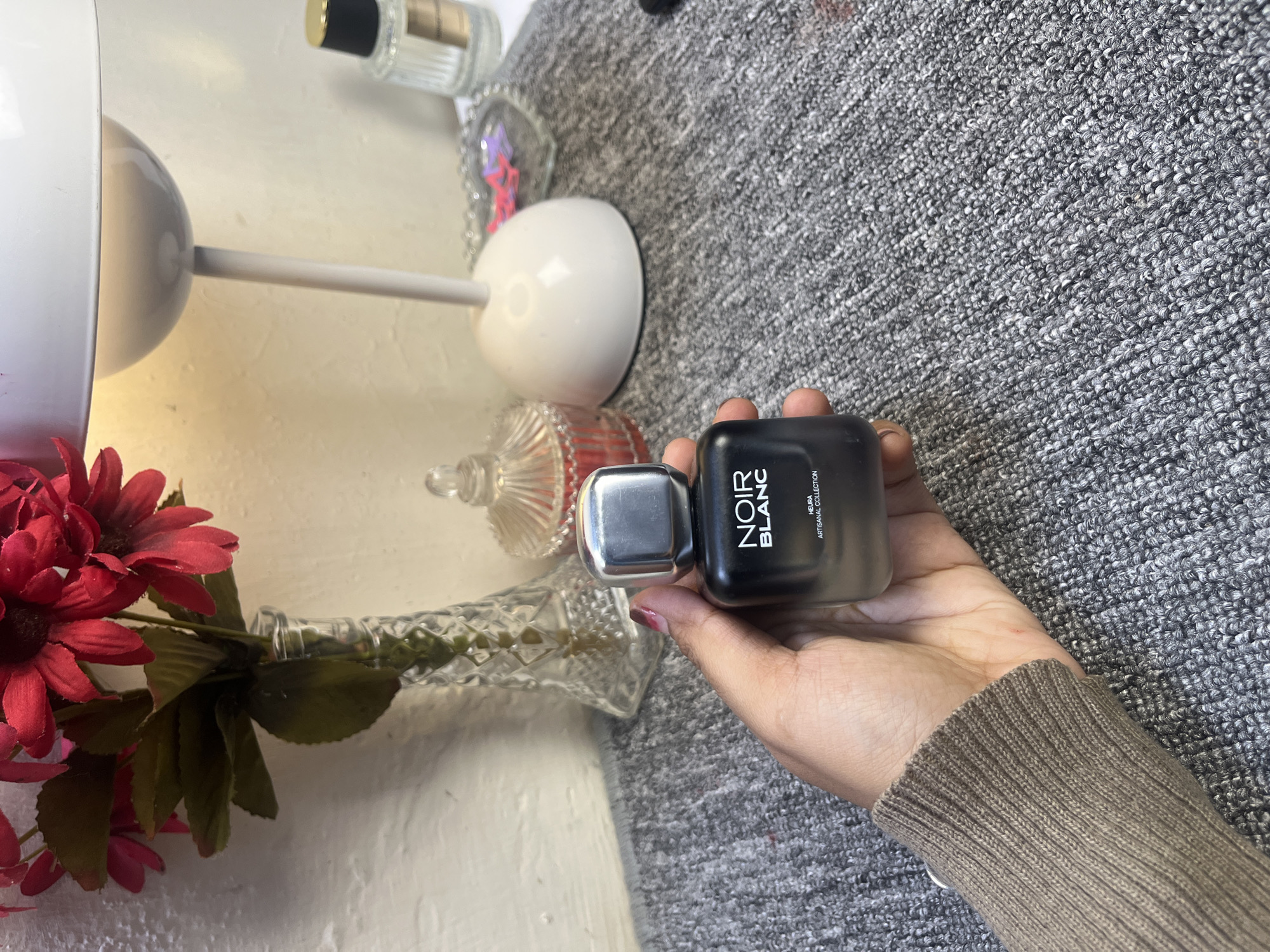 A photograph shows a hand holding a bottle of "Noir Blanc" Heura artisanal collection fragrance, set against a textured gray background. In the background are various decorative items, including a lamp with a white shade, a vase with red flowers, and other bottles. The scene is indoors, and the lighting appears to be from both natural and artificial sources. The image does not provide information to ascertain if this scene is located in Kranggan, Indonesia.