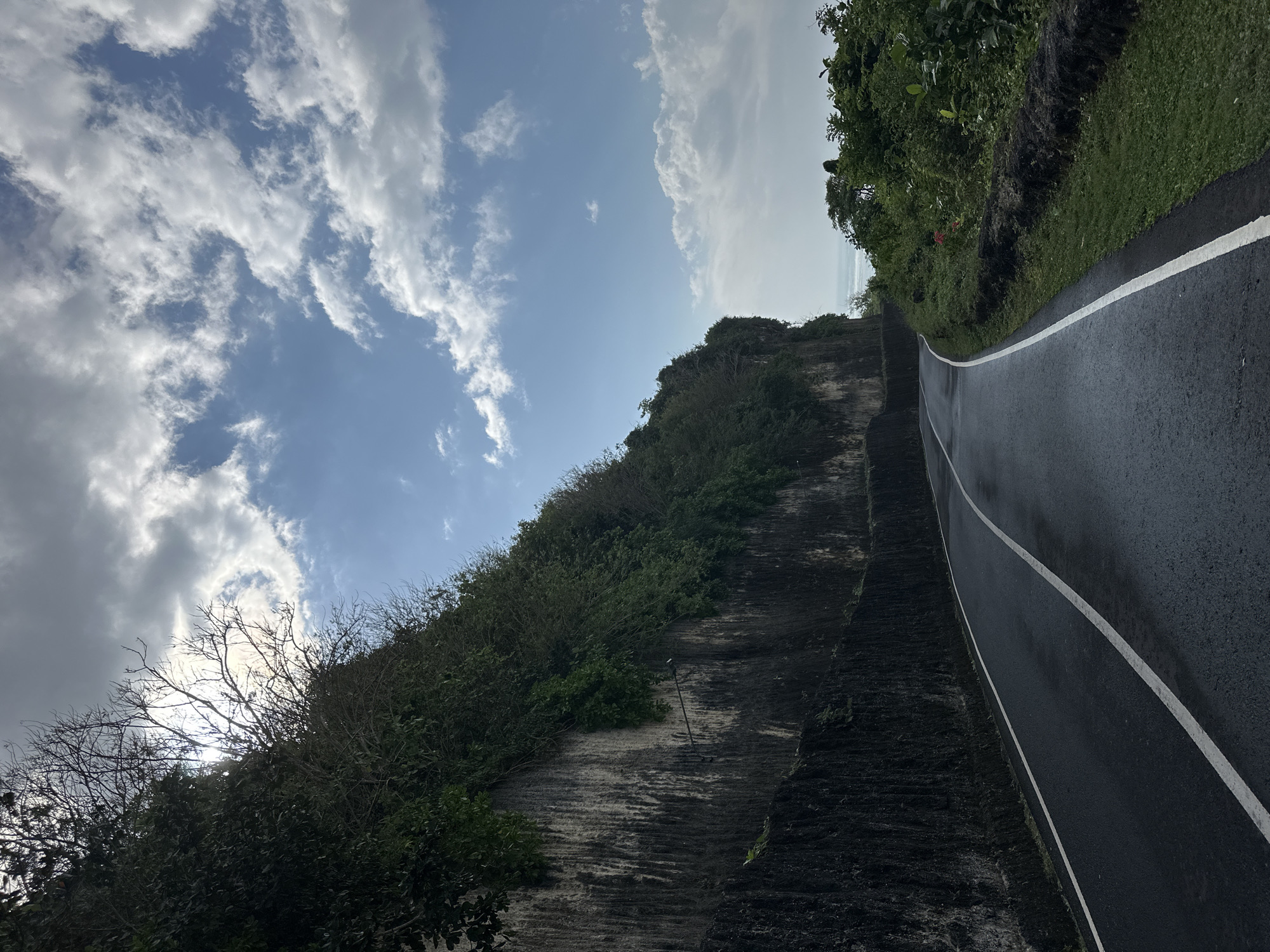 A paved road curves through a tropical landscape. On the left, a sheer, dark rock face rises steeply, covered in dense green foliage and some bare branches. The sun peeks through the clouds behind the branches. On the right, lush green vegetation and trees line the roadside, with a glimpse of the ocean in the distance. The road is dark asphalt with white lane markings, appearing slightly wet. The sky is a mix of bright blue and dramatic, swirling white clouds.