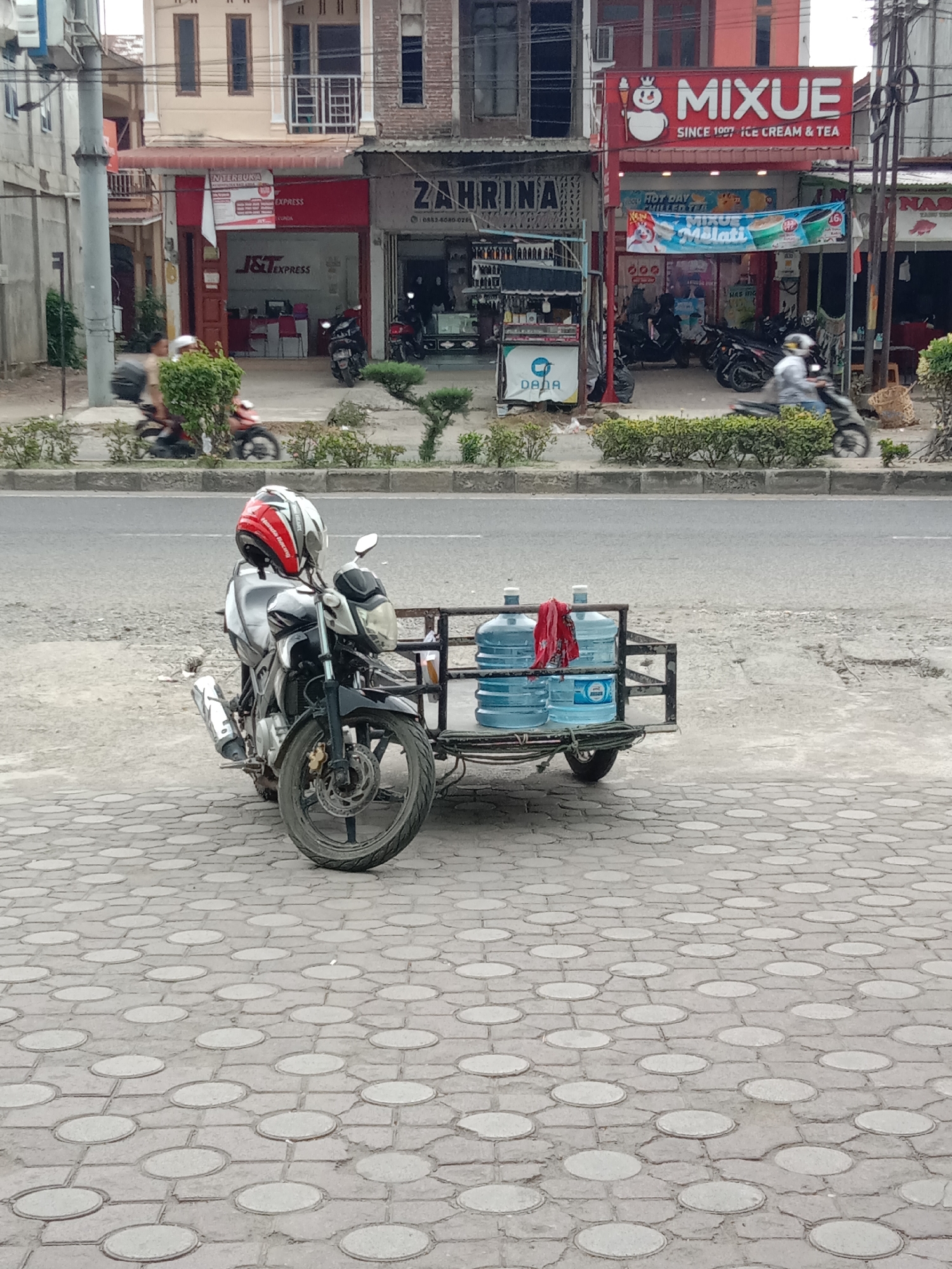 The image shows a black motorcycle with a sidecar parked on a patterned gray brick surface in Lhokseumawe, Indonesia. The sidecar is loaded with four large, clear water bottles and a red cloth. A red and white helmet rests on the motorcycle seat. Across a paved road, several commercial establishments line the street. From left to right, these include "J&T EXPRESS," "ZAHRINA," and "MIXUE SINCE 1997 ICE CREAM & TEA". Other motorbikes are parked near MIXUE and intermittently moving on the road. The architecture consists of multi-story buildings with varied facades. The environment includes street-side greenery planted along the median.